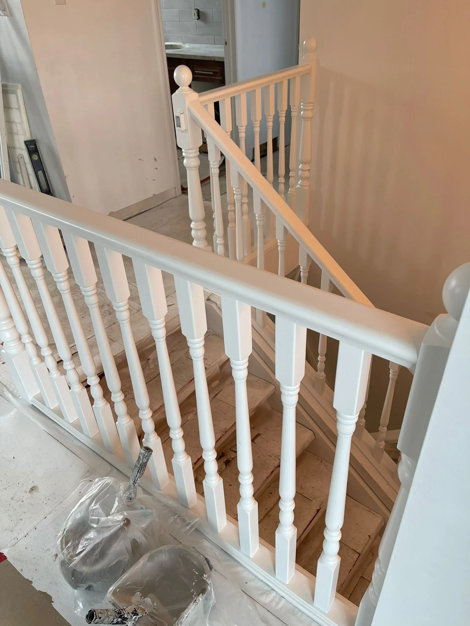 Interior view of a staircase with white wooden balustrades, under renovation. There is a plastic sheet on the floor with a paint roller, and a partially painted staircase.