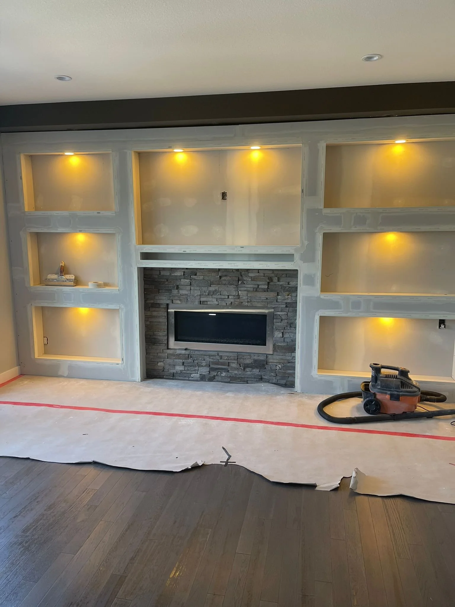 Living room area under renovation with built-in shelves, a stone fireplace, and a partially finished wall with recessed lighting, with flooring and construction tools visible.