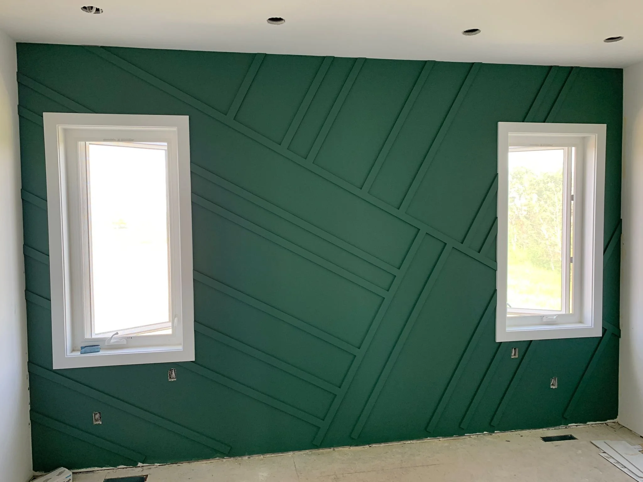 Interior view of a wall with geometric wood paneling painted green, with two rectangular window openings showing outdoor greenery, and several electrical outlets and fixtures not yet installed.