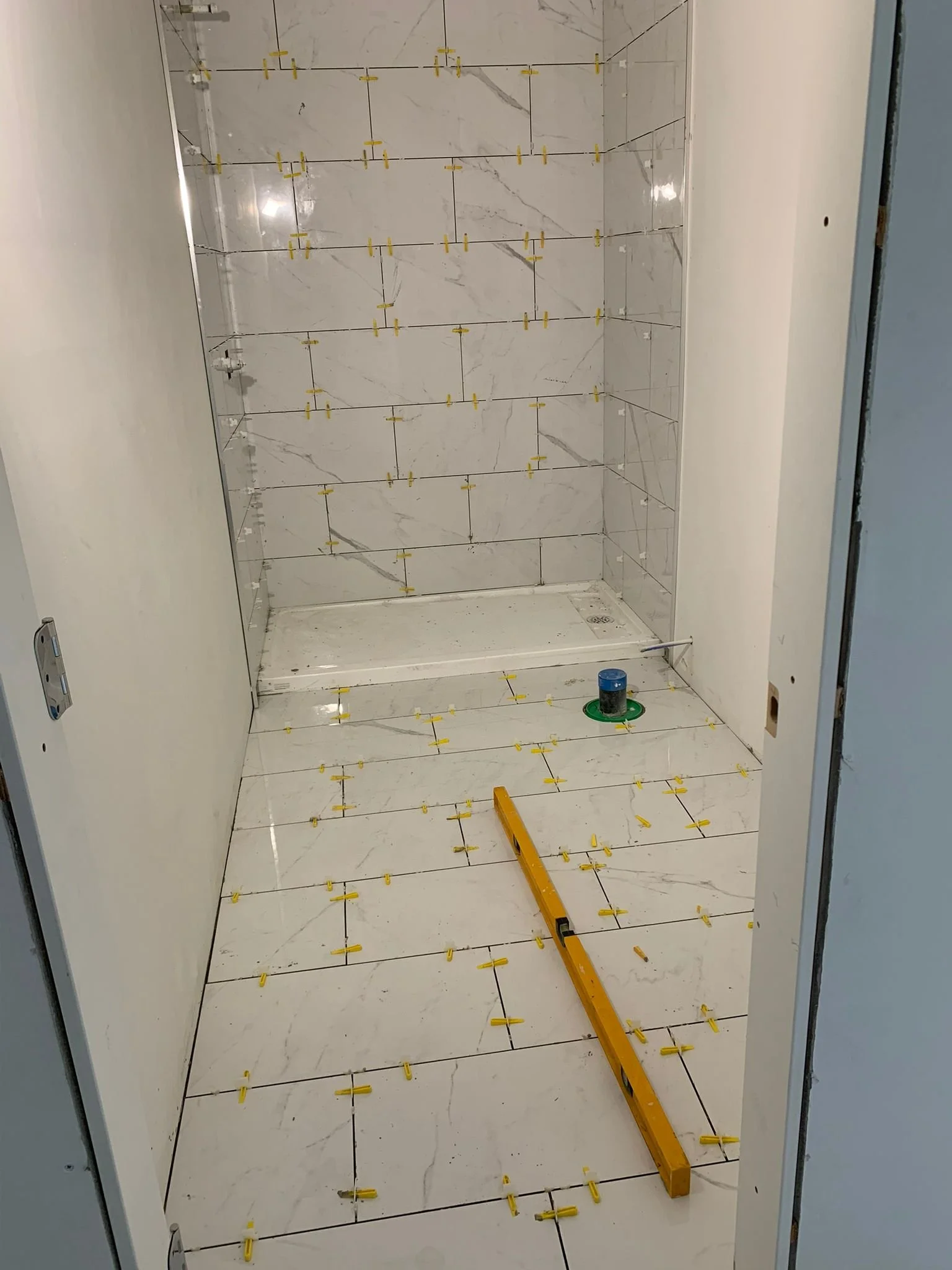 Bathroom with marble-like tiled walls and floor, undergoing tiling work with yellow spacers, a level tool, and a drain in the shower area.