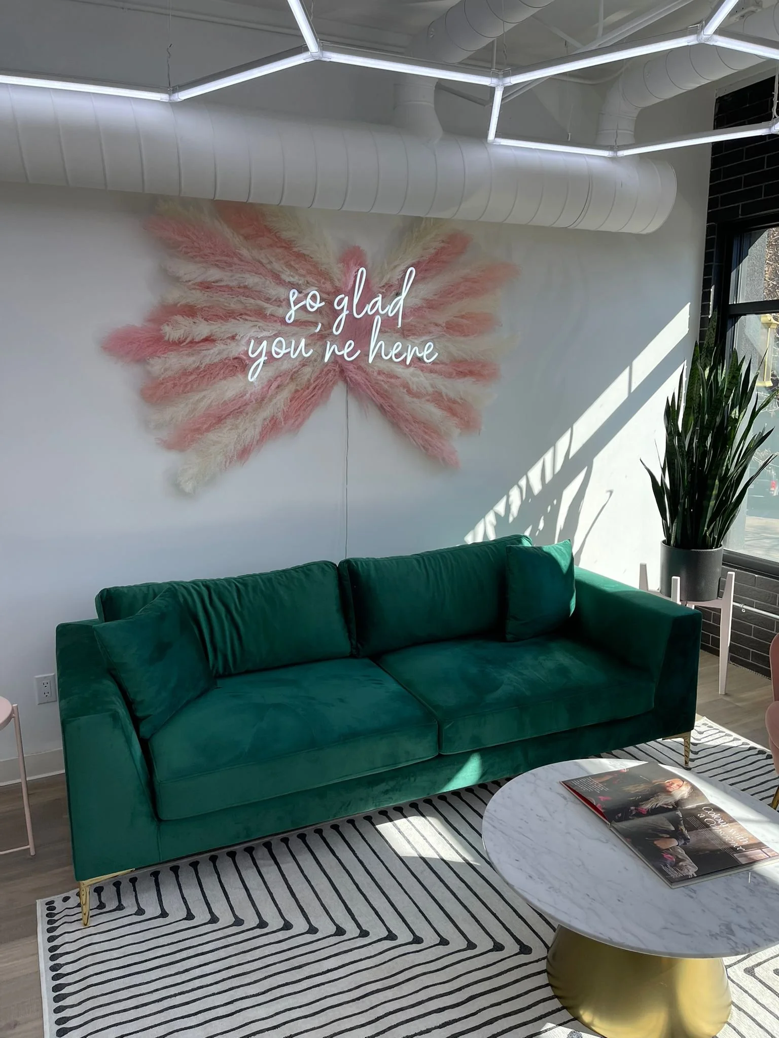 Modern interior space with a dark green velvet sofa, a white and black striped rug, a round white marble coffee table with magazines, a large potted plant, and a decorative wall with pink and beige feather-like art and a neon sign that says 'so glad 