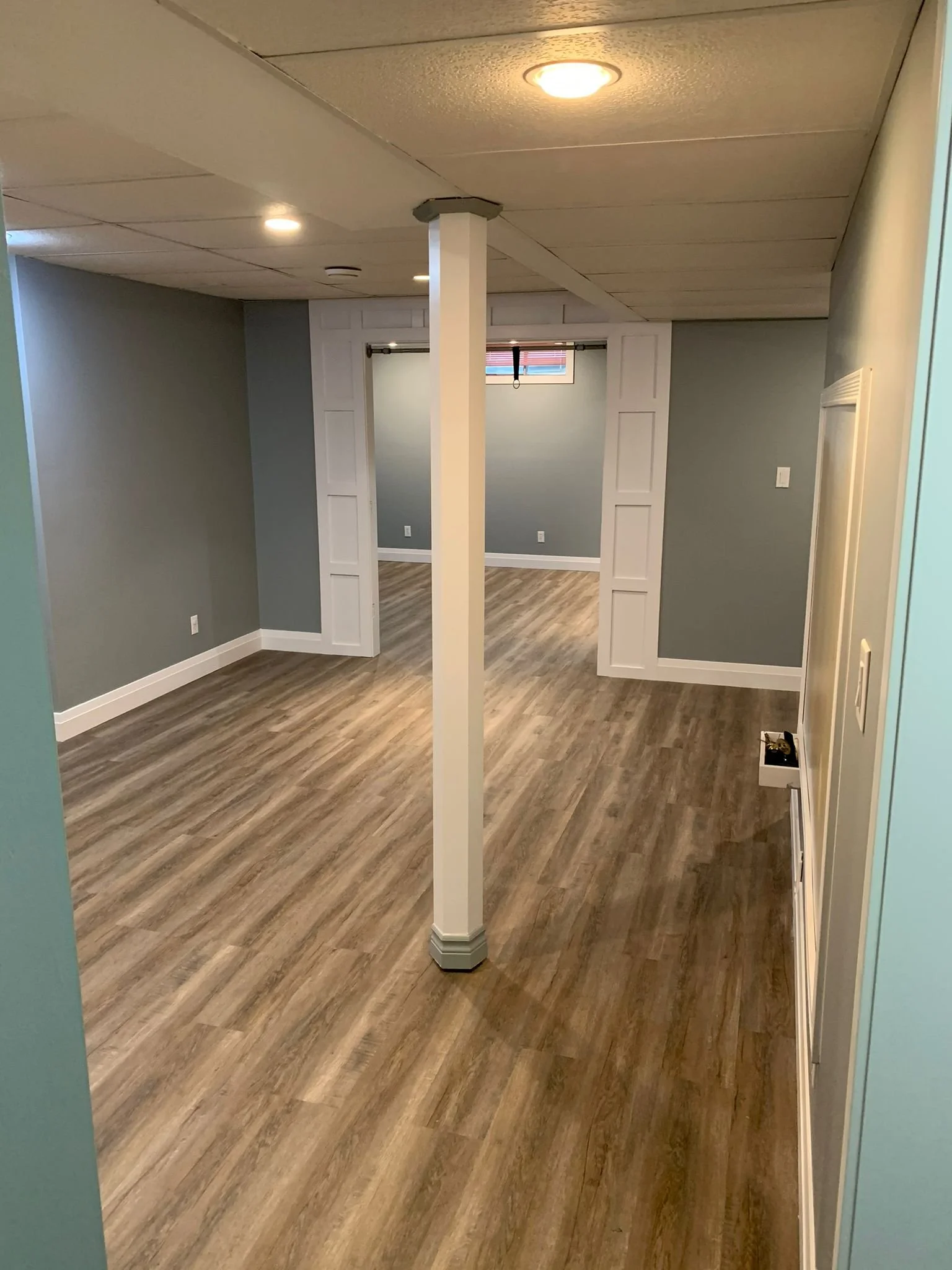 Empty room with wood flooring, gray walls, a white column, small upper window, ceiling lights, and a small object on the floor near the wall.