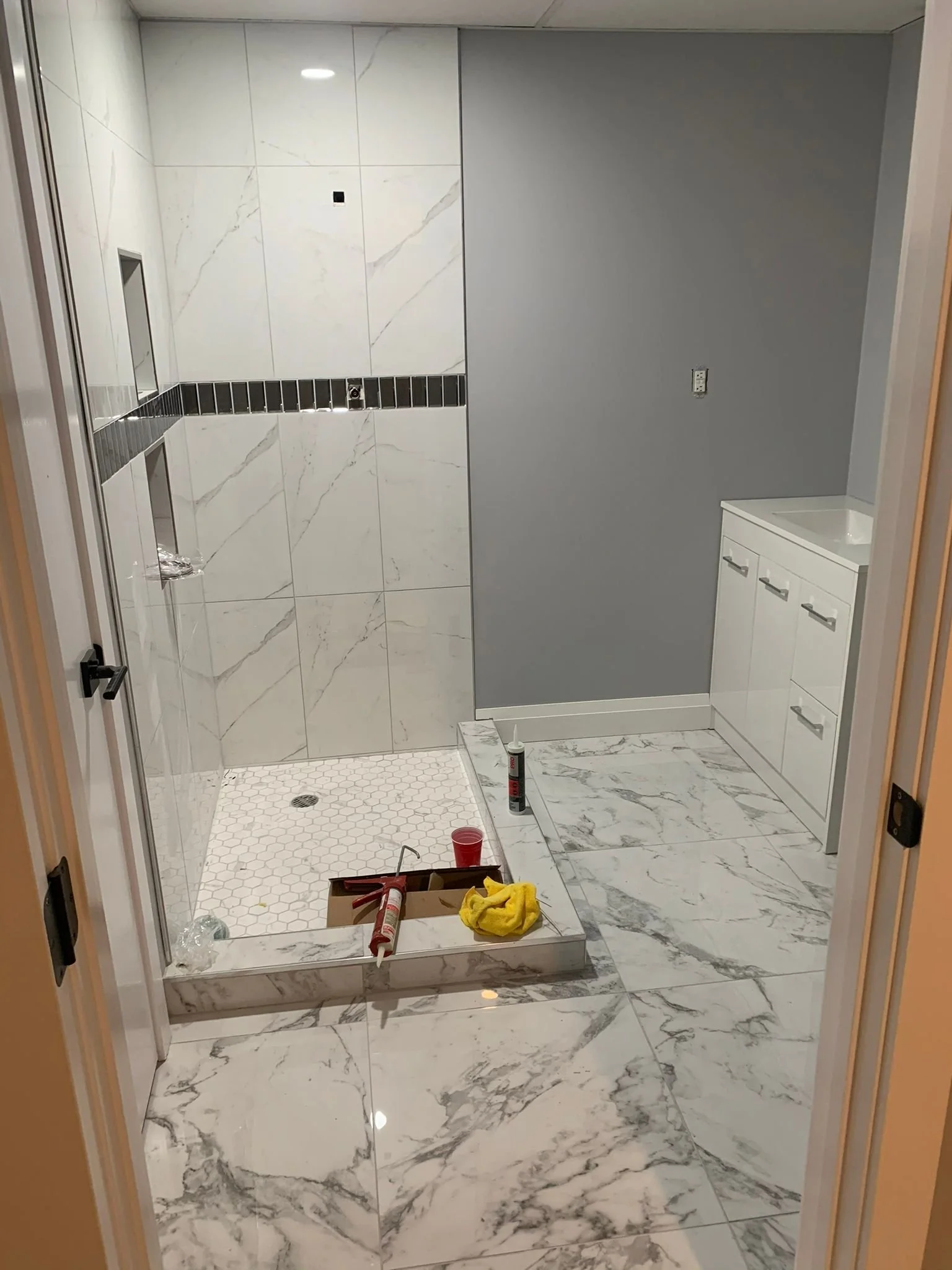 A bathroom under renovation with white marble floors and walls, a shower area with a hexagonal tile floor, a silver and black strip tile border on the wall, a built-in wall niche, and a small white vanity with drawers. Construction tools and supplies