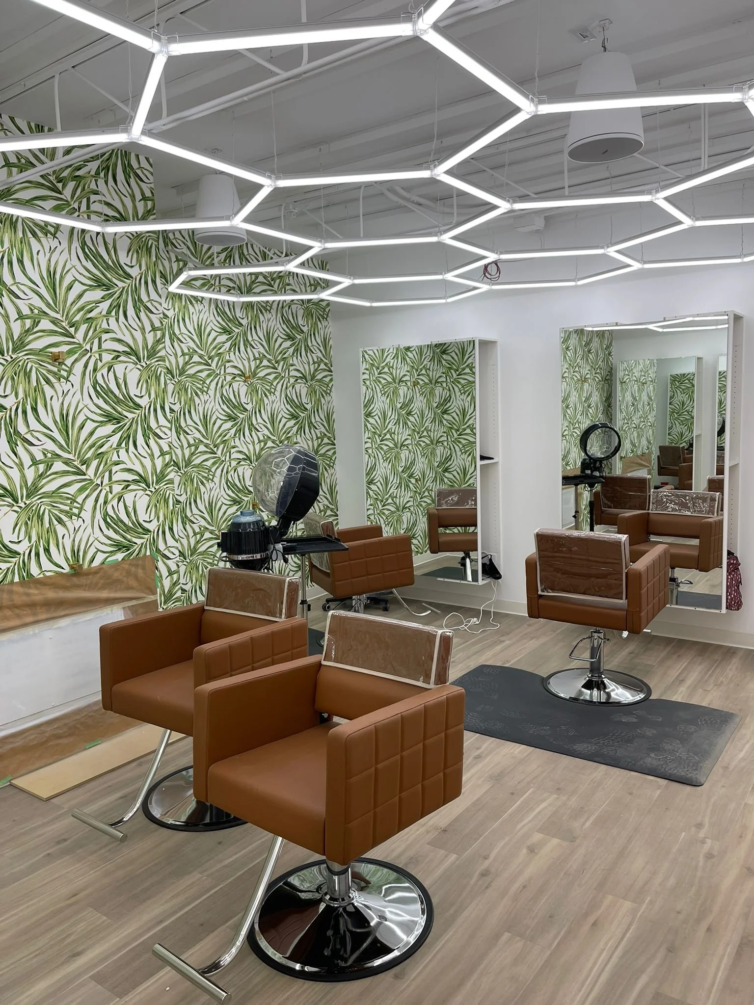 A modern salon with brown leather barber chairs, large mirrors, and a patterned green leafy wallpaper. Overhead, there are white geometric LED light fixtures and some hair styling equipment visible.