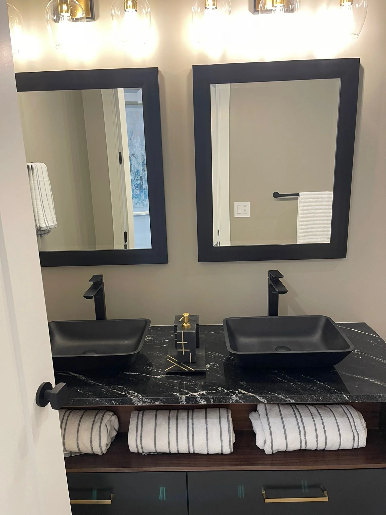 A modern bathroom vanity with two black vessel sinks on a black marble countertop, with black faucets. Two large, rectangular mirrors with black frames are mounted above the sinks. Towels are rolled underneath the counter. The wall behind has wall-mo