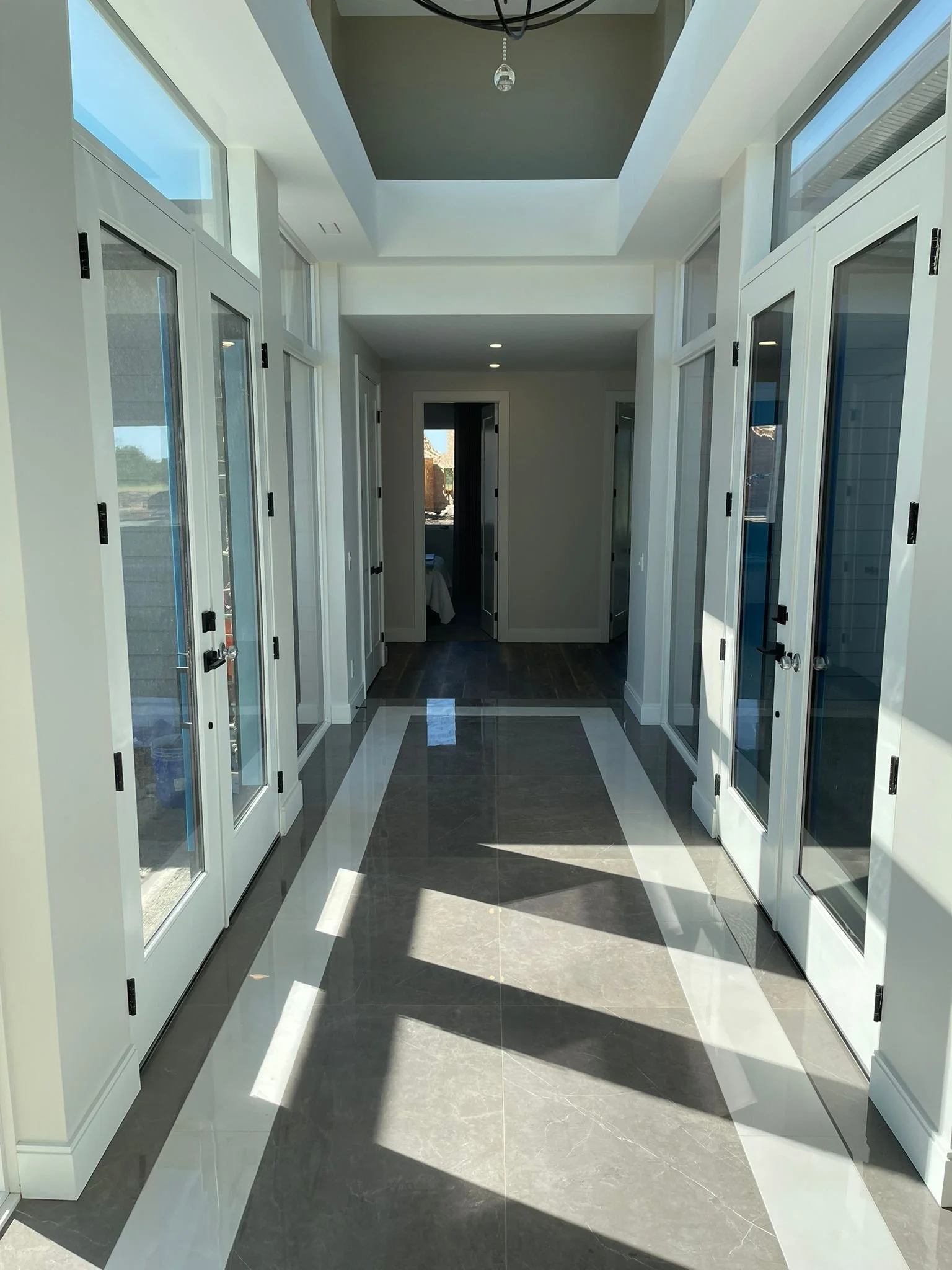 A bright hallway with large glass doors on both sides, leading to the outside, and a view of a room at the end. Sunlight casts shadows on the shiny tiled floor.