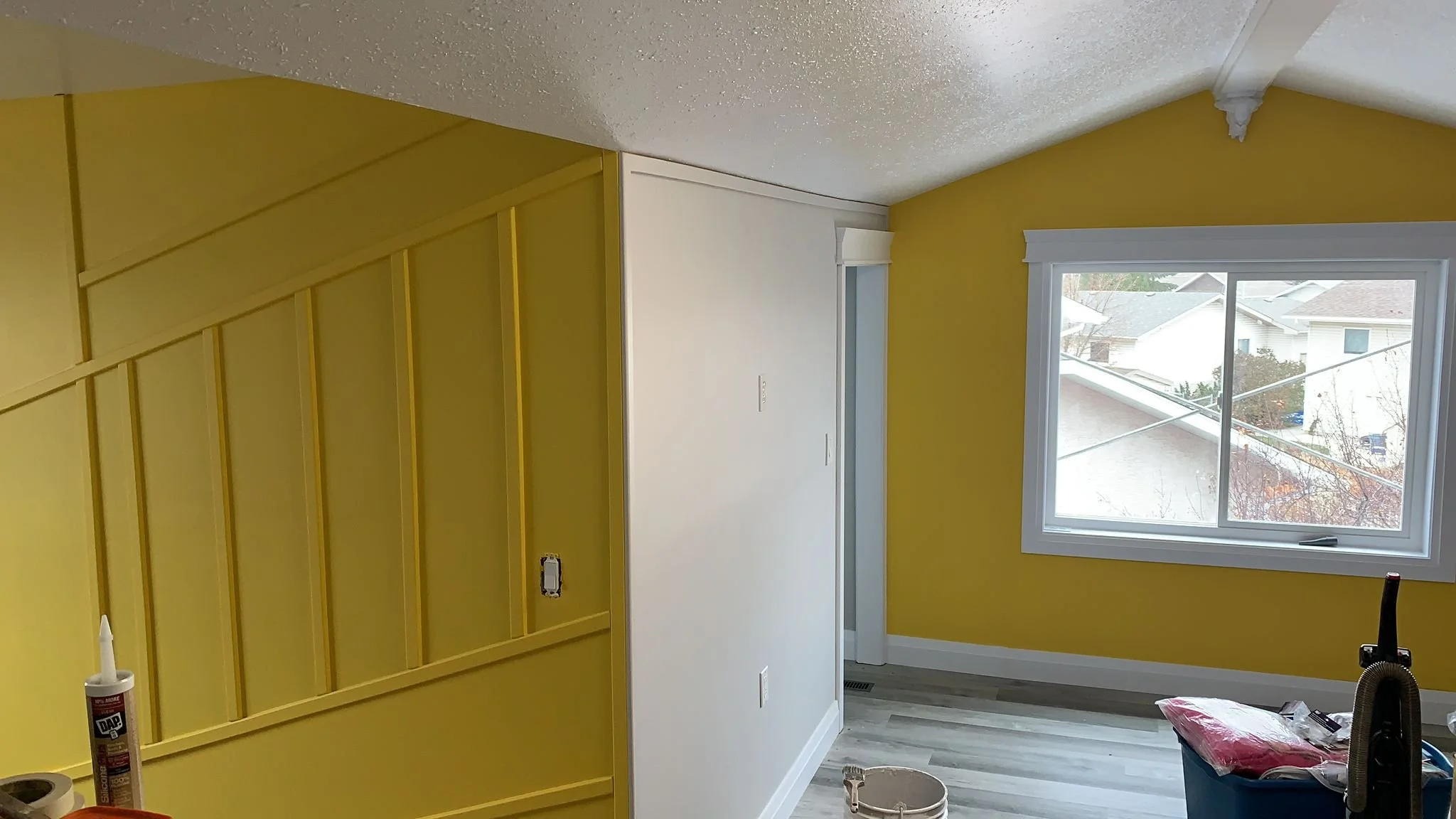 A room with partially painted yellow walls, including areas around a window and part of a ceiling. There are painting supplies and tools such as a caulk tube, a bucket, and miscellaneous items on the floor.