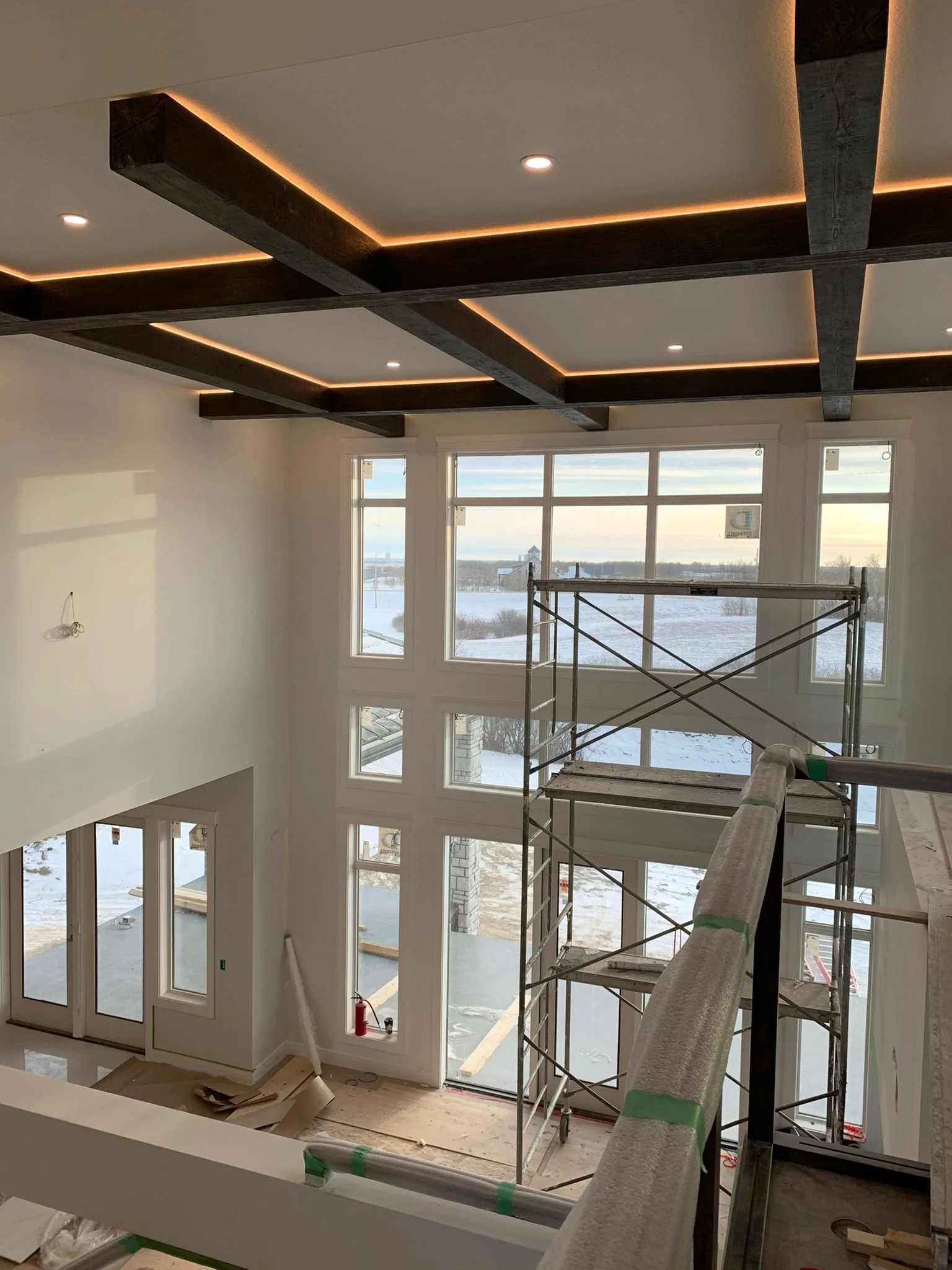 Interior of a house under construction with a modern ceiling design featuring wooden beams and LED strip lighting, large windows showing a snowy landscape outside, and scaffolding inside.