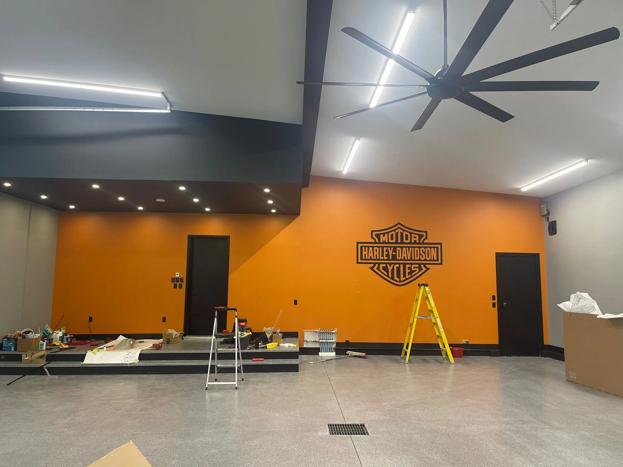 An interior space under construction or renovation with a large orange wall featuring a Harley-Davidson logo, a ceiling fan, fluorescent lighting, and construction tools and supplies scattered on the floor.