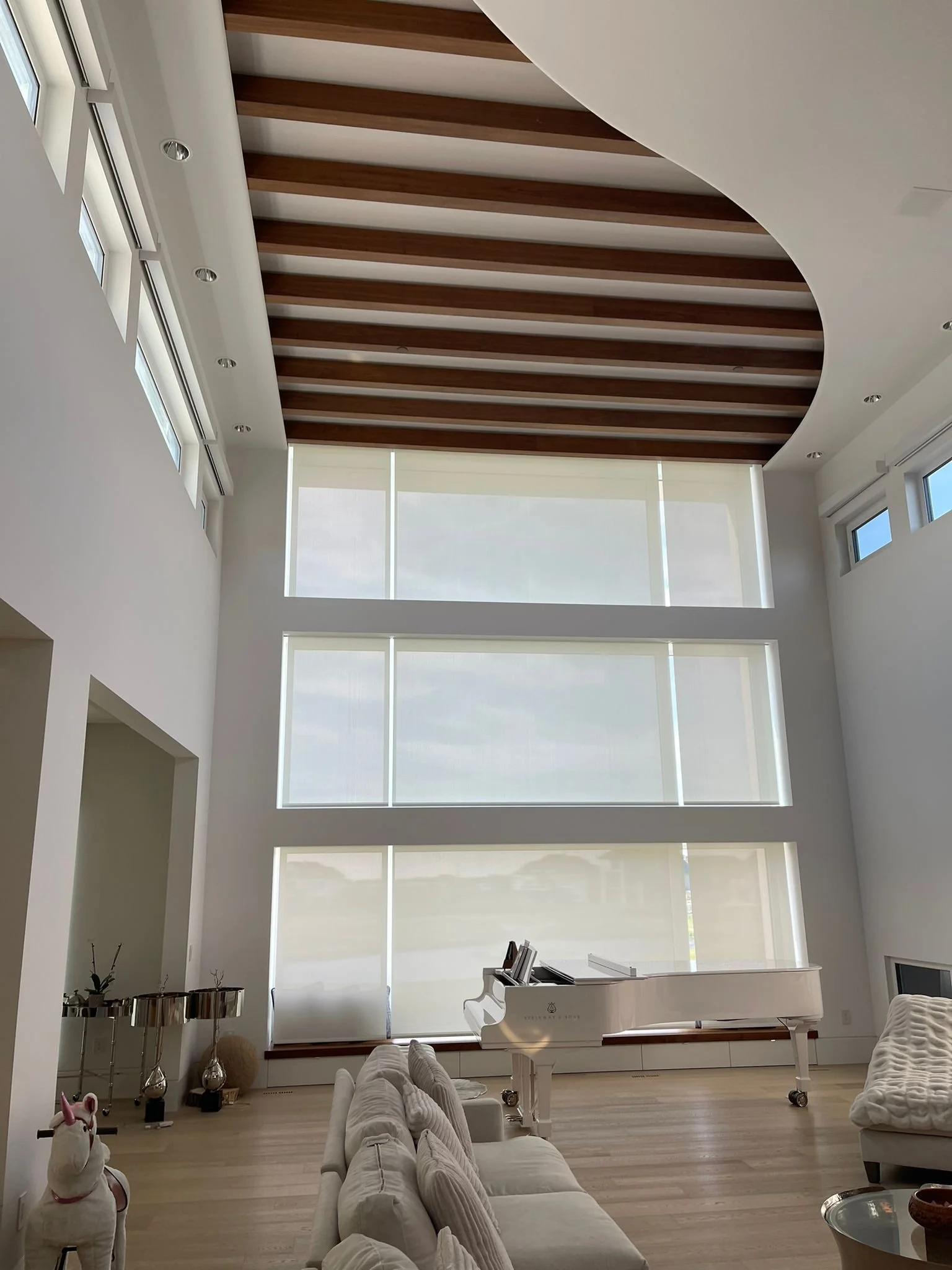 Bright living room with white walls and a lofty ceiling featuring wooden beams. Large windows with white shades let in natural light. Elegant white sofa and armchair arranged around a white grand piano. Decorative items include a unicorn plush toy, v