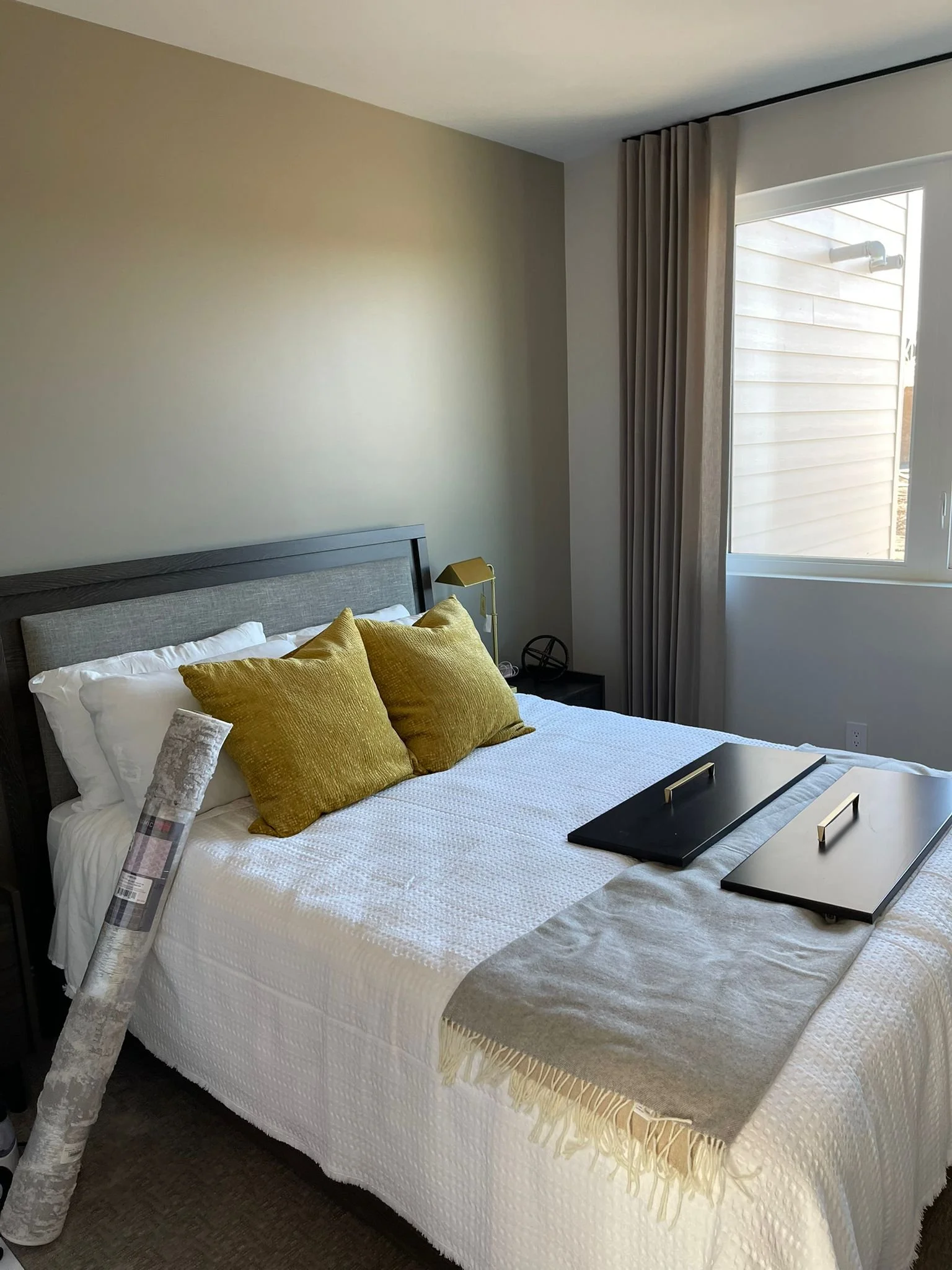 A bedroom with a bed, yellow pillows, a nightstand, a yellow lamp, a window with beige curtains, and a view of the outside.