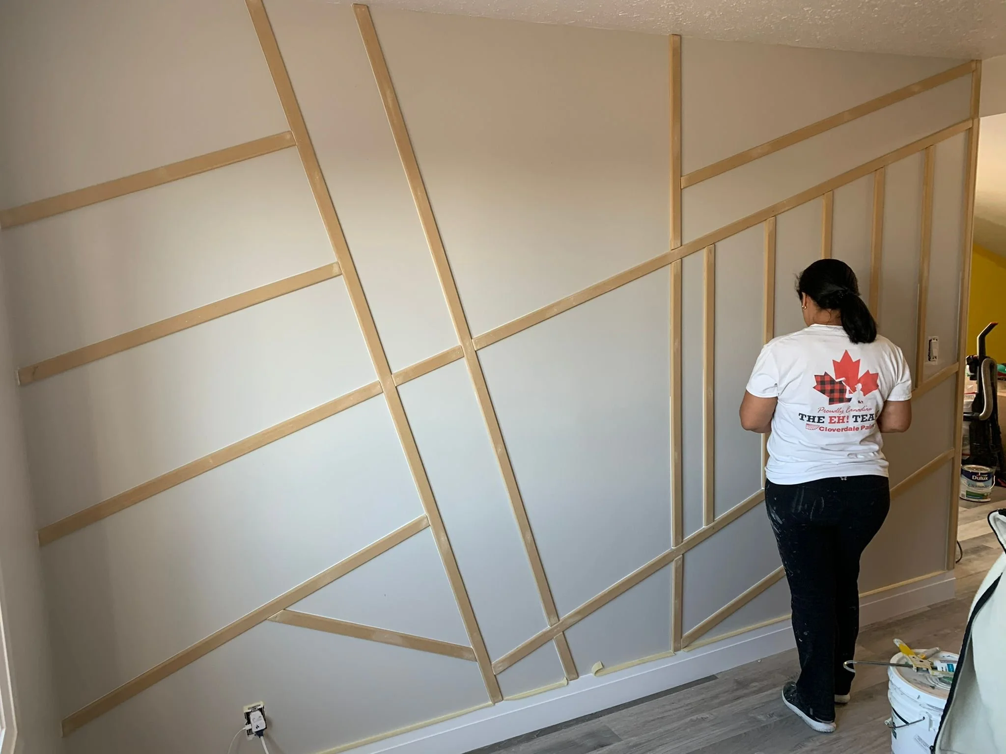 A woman in a white t-shirt with a maple leaf and text on the back is building a wooden frame wall in a room. The wall has vertical and angled horizontal wooden pieces attached to a white wall.