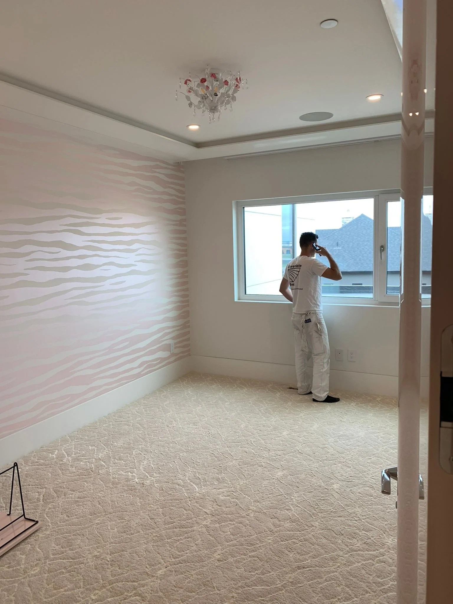 A person standing in a bright, empty room with beige carpet, white walls, and a window, talking on a phone. There is a pink and white patterned wallpaper on one wall, recessed ceiling lights, and a decorative chandelier.
