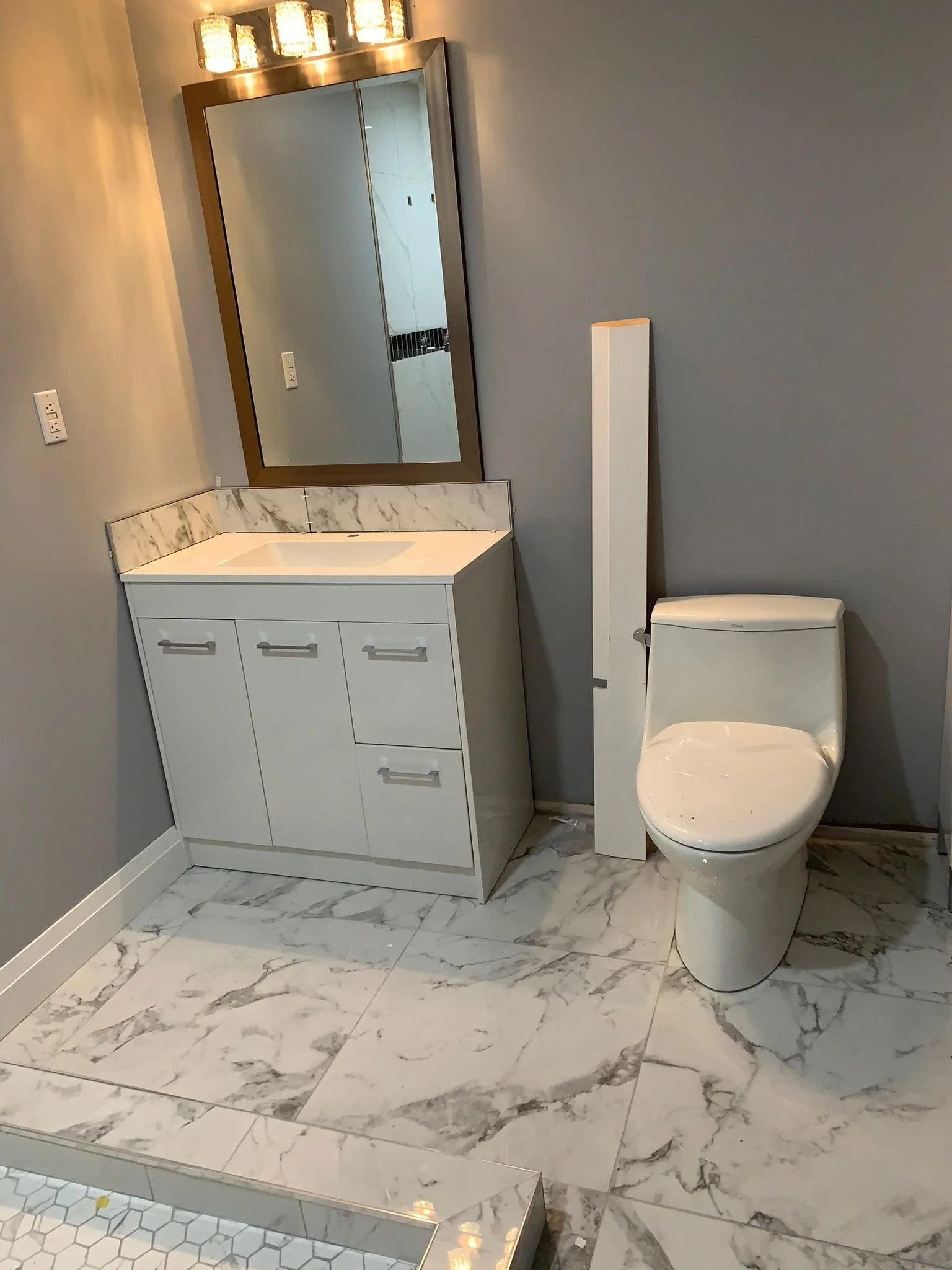 A modern bathroom with white marble tile floor, a white vanity with marble countertop, a large mirror, wall sconces, a white toilet, and gray painted walls.