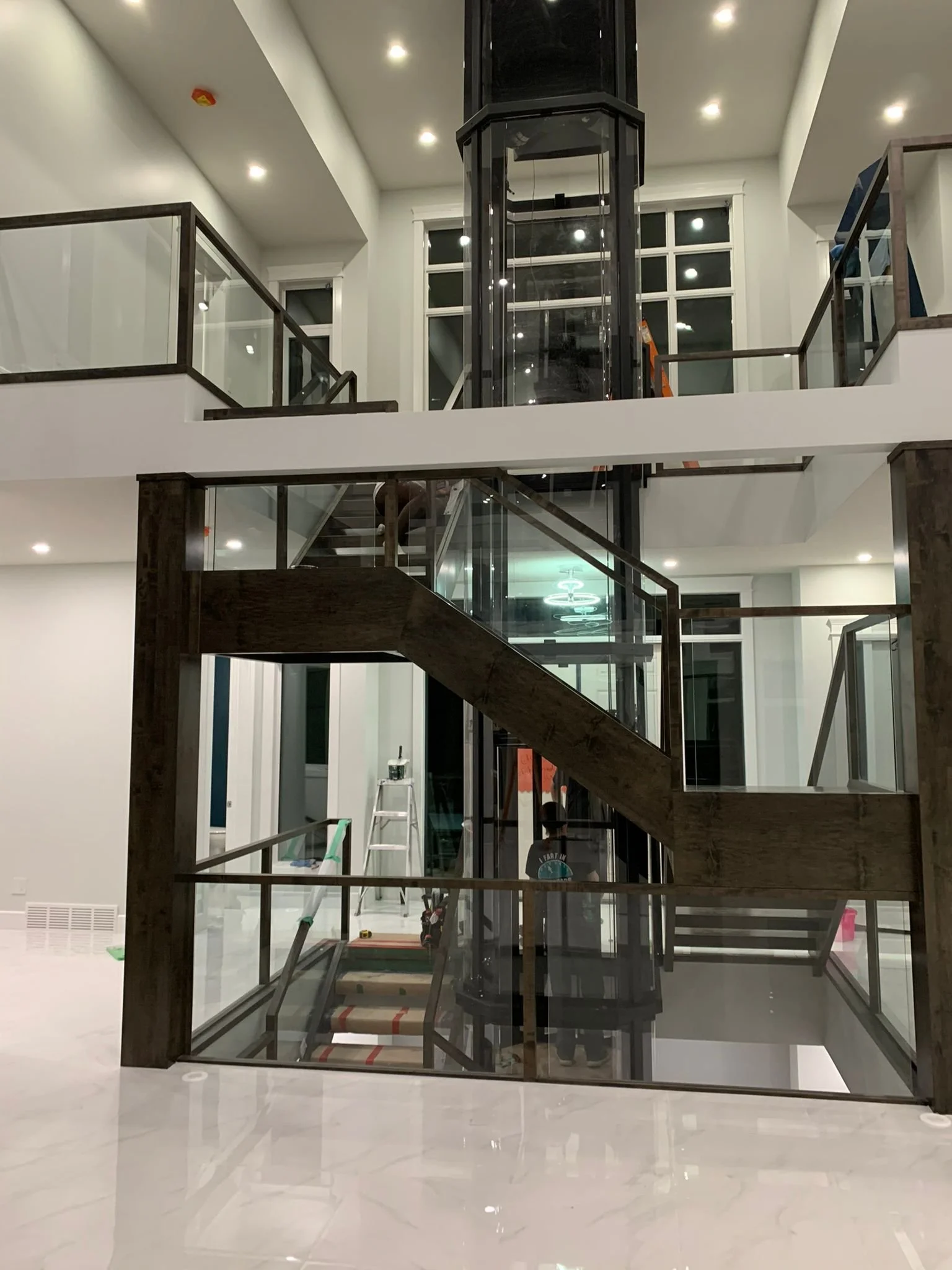 Interior view of a modern building under construction, featuring a glass elevator shaft, stairs with glass railings, white walls, and ceiling lights.