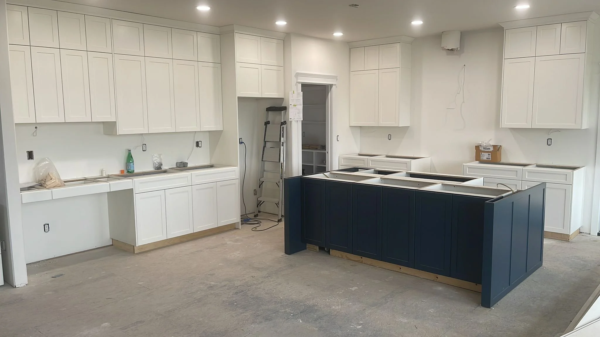 A kitchen under construction with white upper and lower cabinets, a dark blue island, and unfinished flooring.