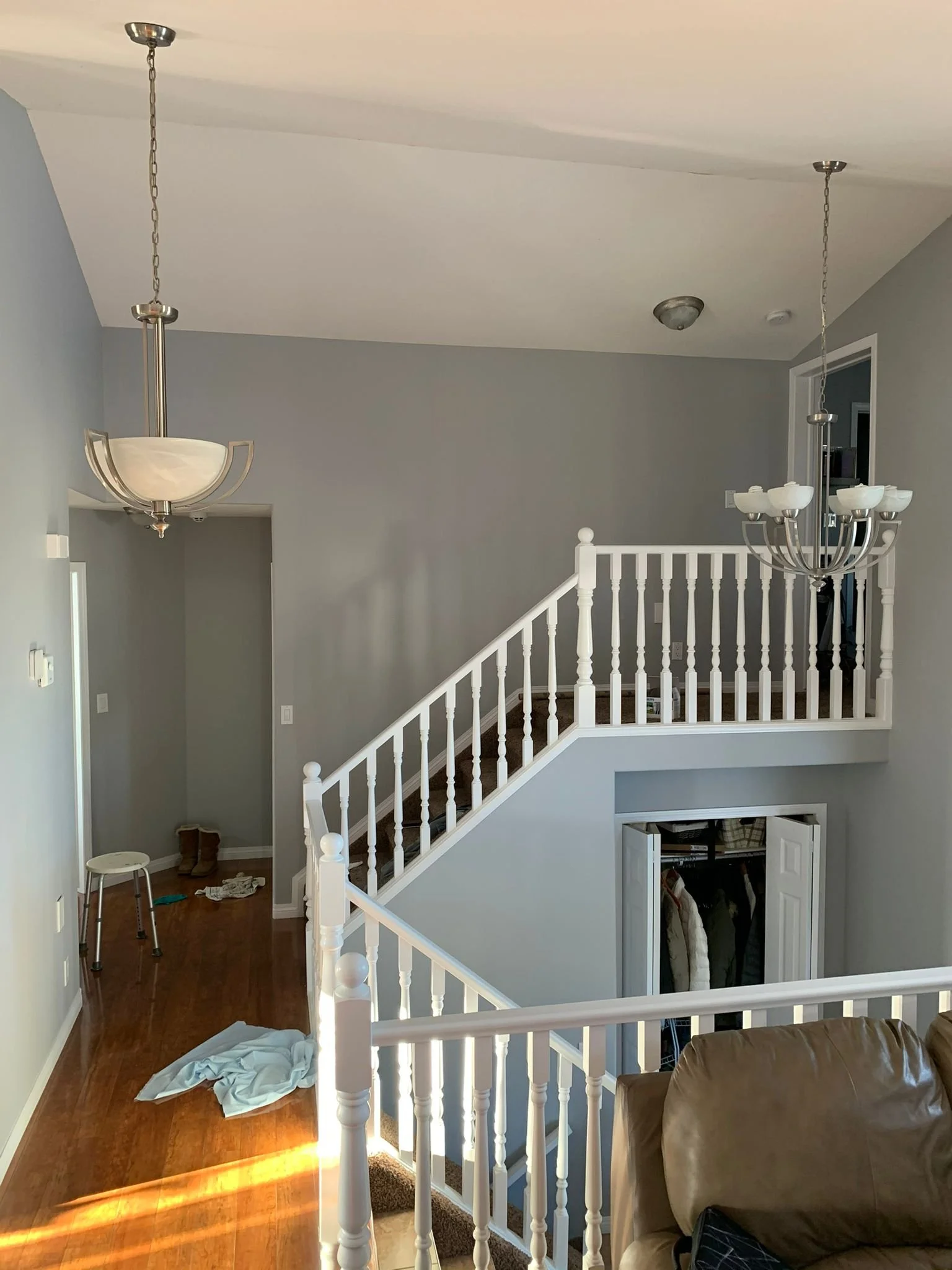 View of a house's staircase with a chandelier hanging from the ceiling. There's a brown leather chair in the foreground, and a closet under the staircase with clothes hanging inside. The floor is wooden, and there are some items on the ground near th
