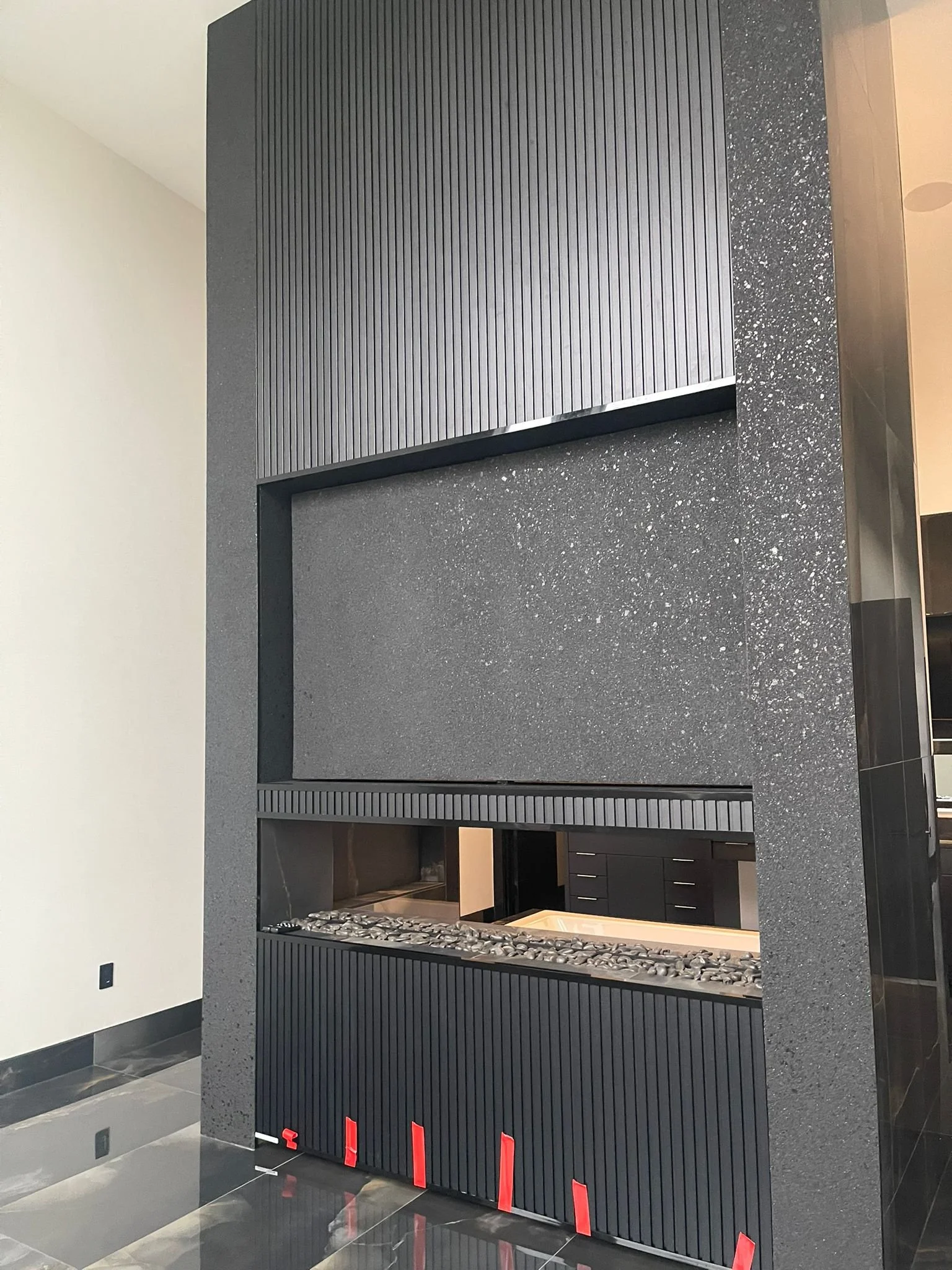 Modern fireplace with a black wooden frame and black pebbles in the firebox, set against a speckled black wall in a contemporary interior.