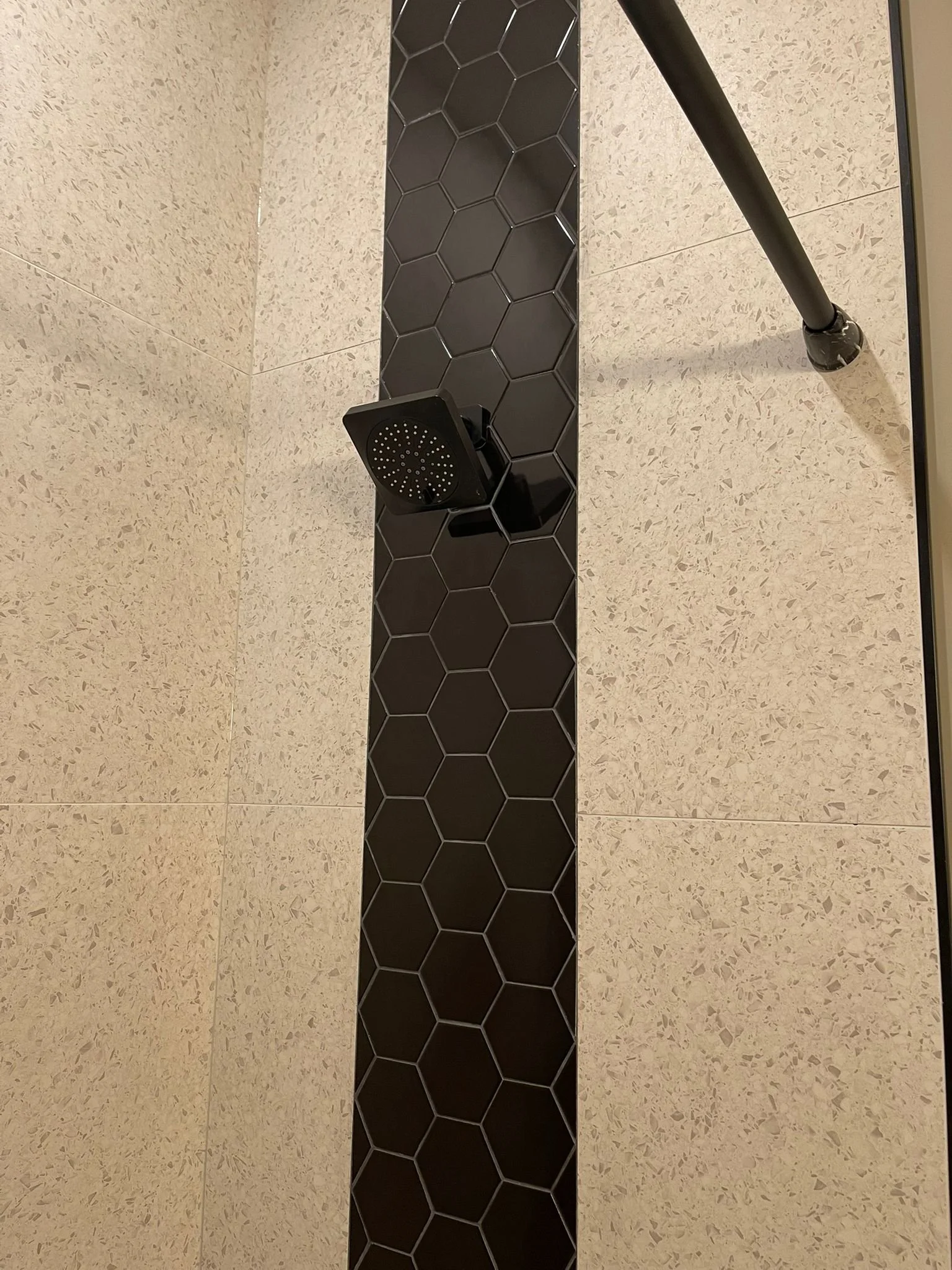 A showerhead attached to a black hexagonal tile wall in a bathroom with beige tiled flooring.