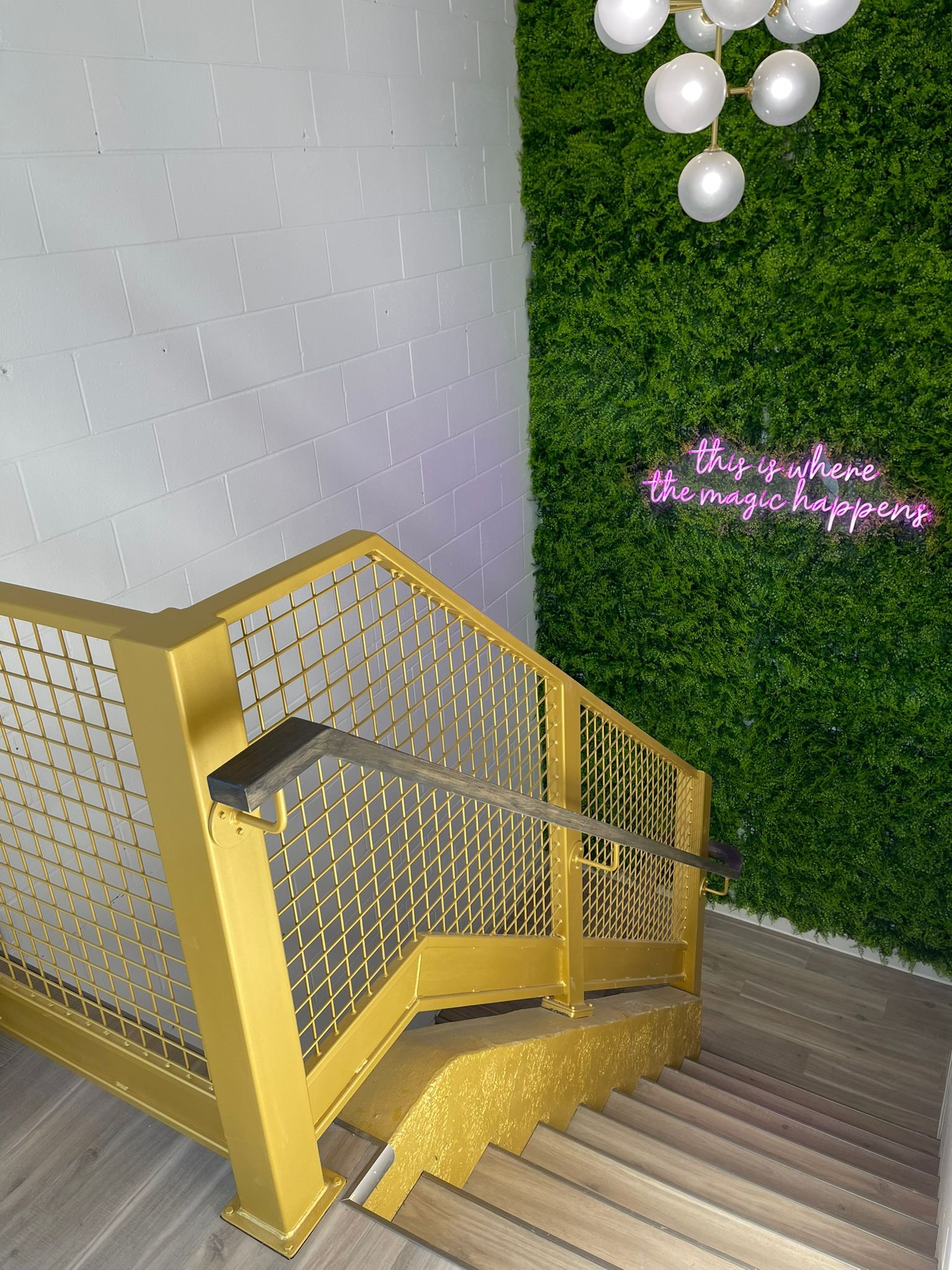 Yellow staircase with metal railings and wooden steps, leading up to a green wall of plants. There is a pink neon sign on the green wall that reads 'this is where the magic happens.' A modern chandelier with spherical bulbs hangs above.