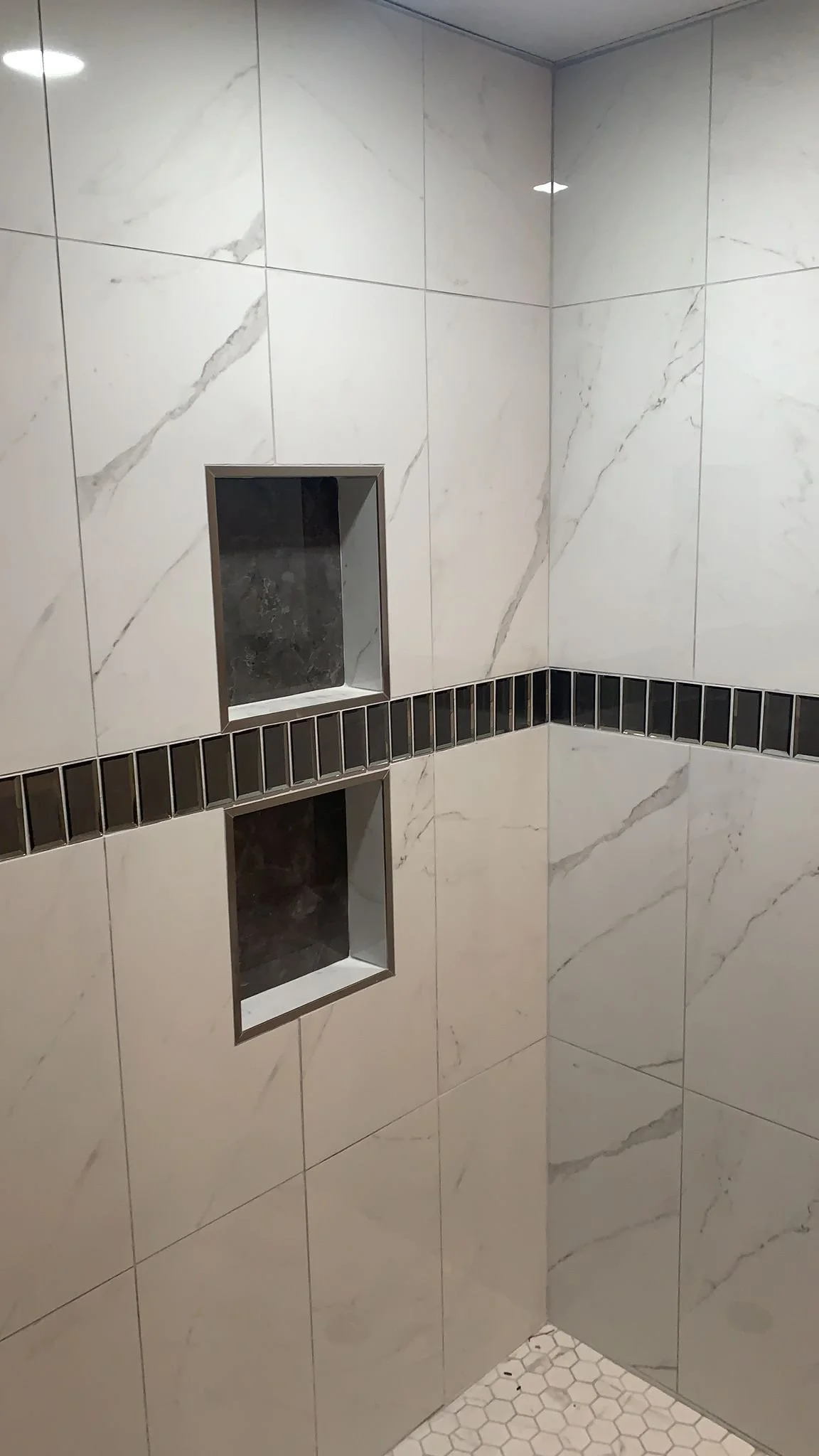 A corner of a shower with white marble wall tiles with gray veins, two recessed shelves with dark tiles, a black border of small rectangular tiles, and a white hexagonal tiled floor.