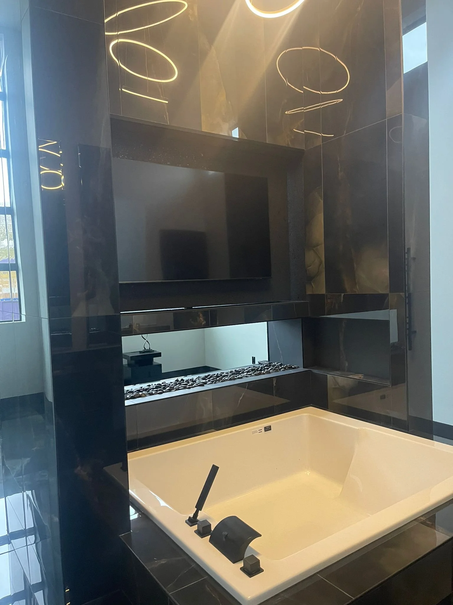 A modern bathroom with a large white bathtub, black tiles, a flat-screen TV mounted on the wall, and ambient lighting fixtures.