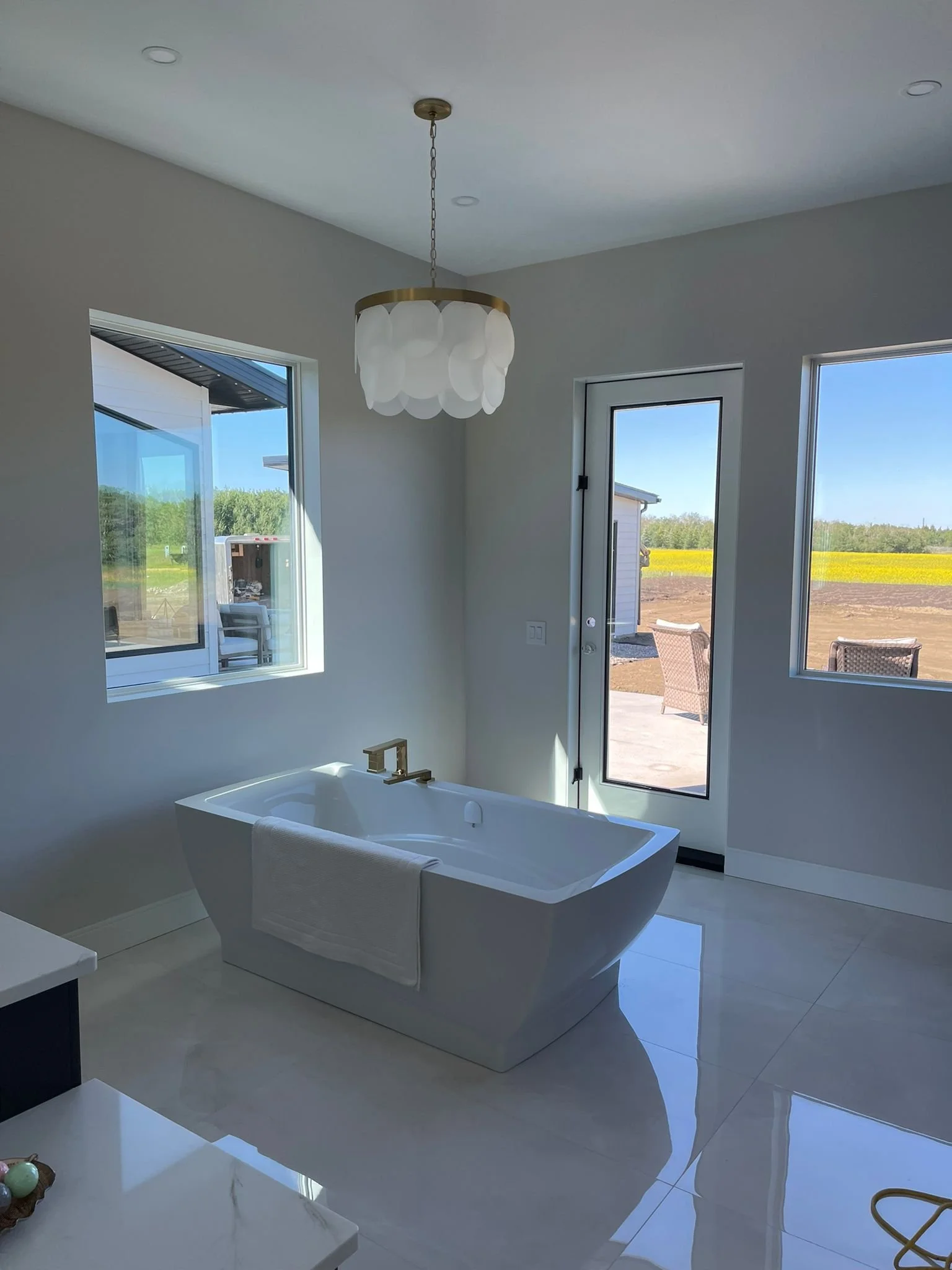 A modern bathroom with a bathtub beneath windows showing a bright outdoor landscape with fields and trees. There is a white towel hanging on the edge of the bathtub, and a light fixture with a gold ring and white decorative panels hanging from the ce