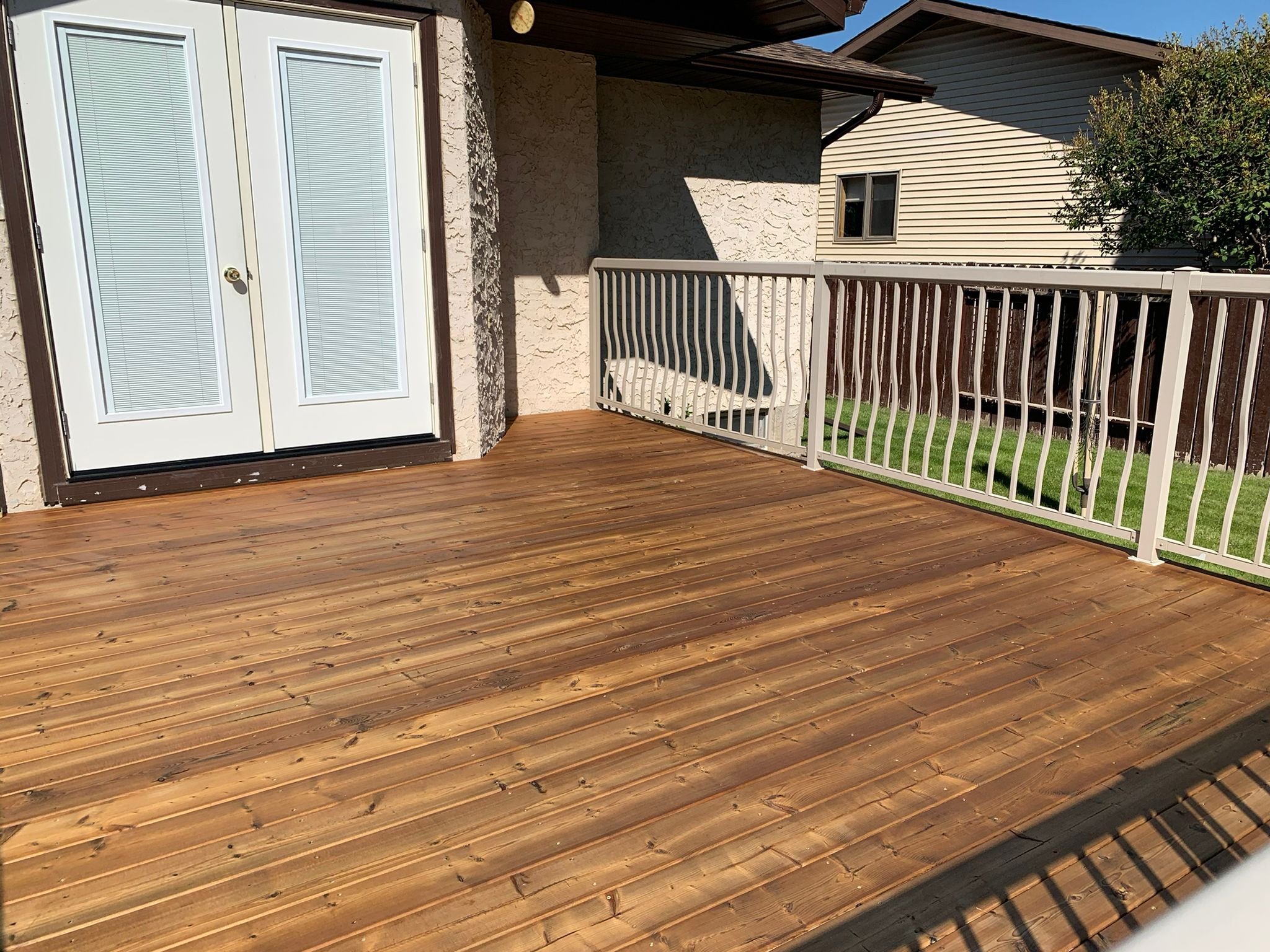 A wooden deck with a railing, white double doors with glass panes, a small attached porch wall, and neighboring houses with siding and a small window, under a clear sky.