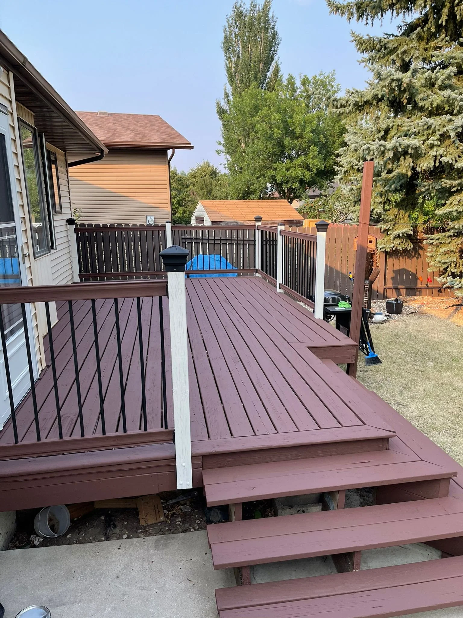 A backyard deck with new wooden reddish-brown floorboards and black and white railing posts, with stairs leading down to a concrete patio and a lawn area.