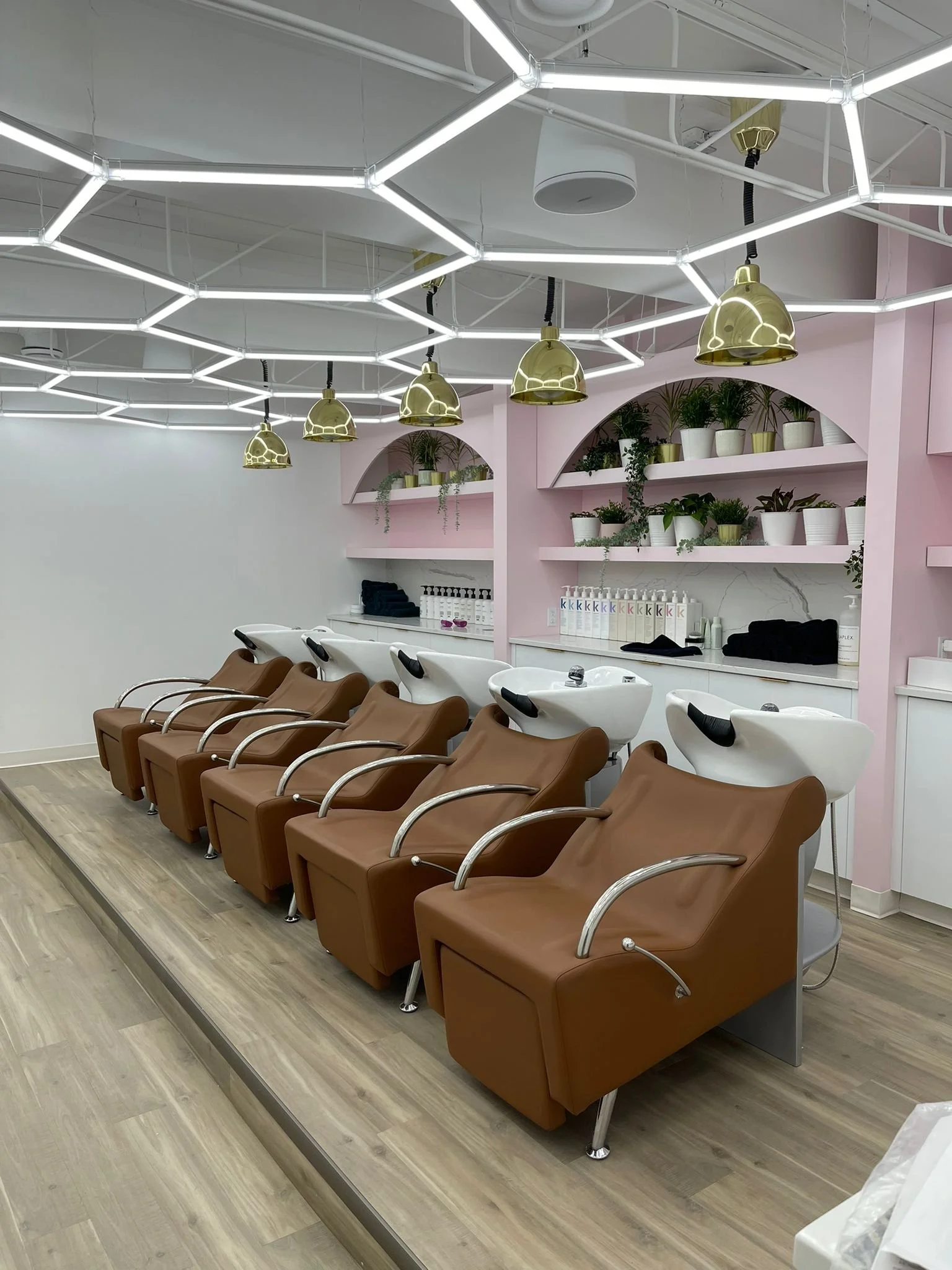 Hair washing stations with brown chairs and white sinks in a salon, featuring a pink wall with shelves holding green and white plants, and a modern ceiling light fixture.