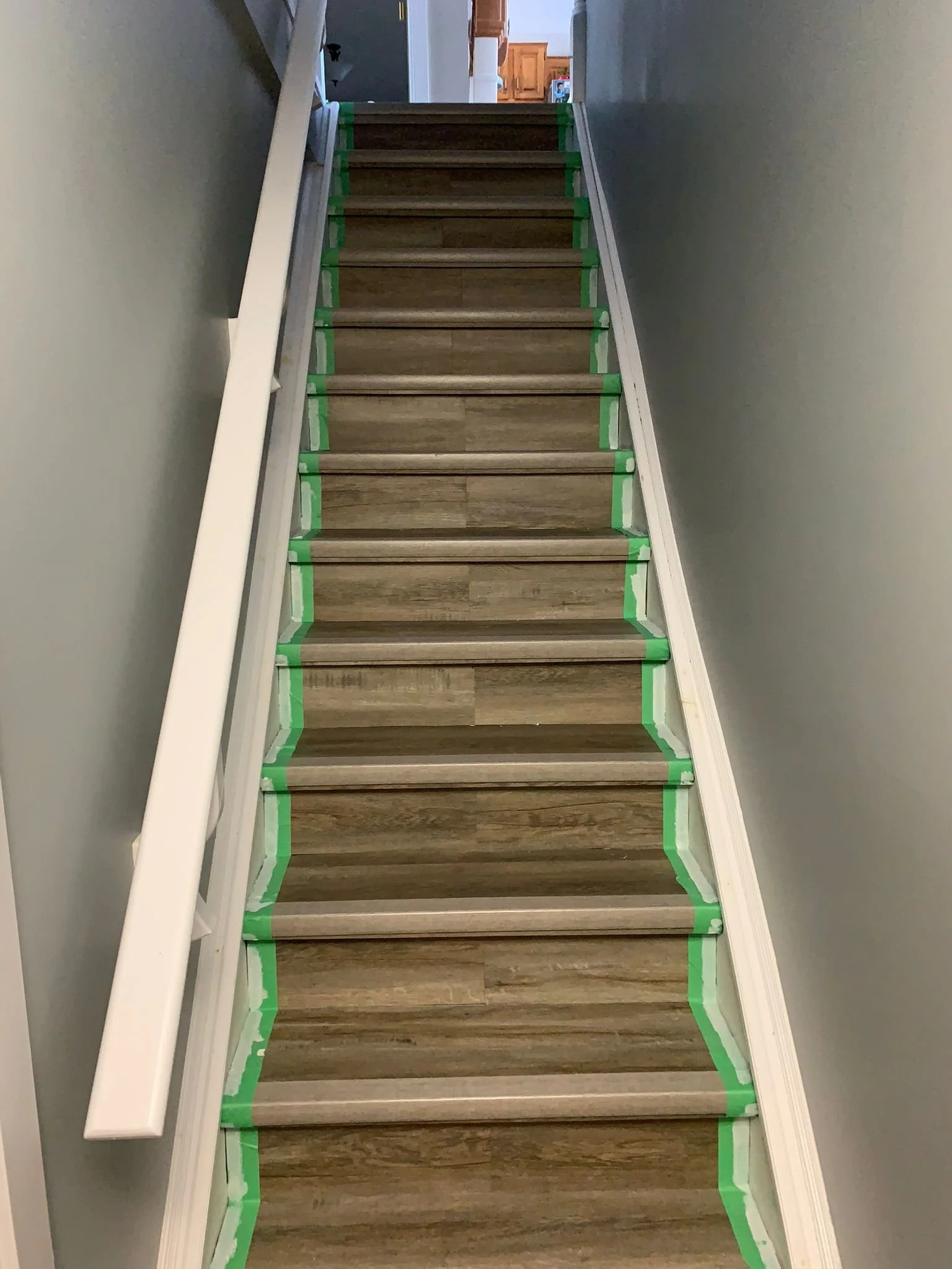 A staircase in a house with new wood flooring, lined with green painter's tape along the edges of each step. The staircase has a white handrail on the left side, and the walls are painted gray. At the top of the stairs, the doorway opens into a kitch