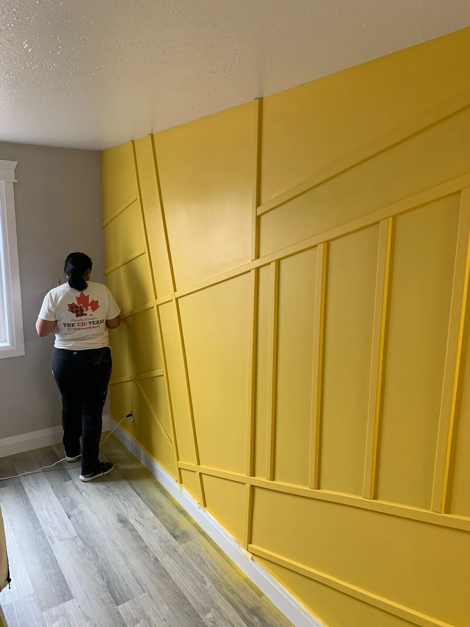 Person standing near a yellow wall with angled panels, working on the wall in a room with light-colored patterned flooring and a white window frame.