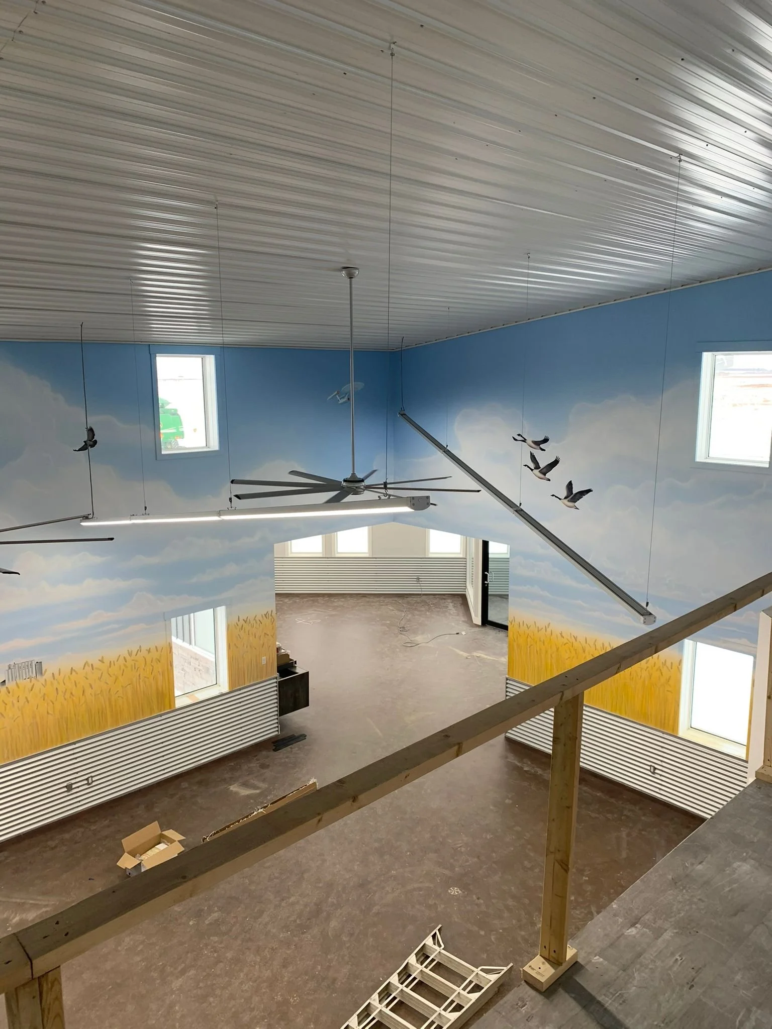 Interior of a building under construction with a high ceiling, fans, large windows, blue cloud mural, and an elevated wooden platform.