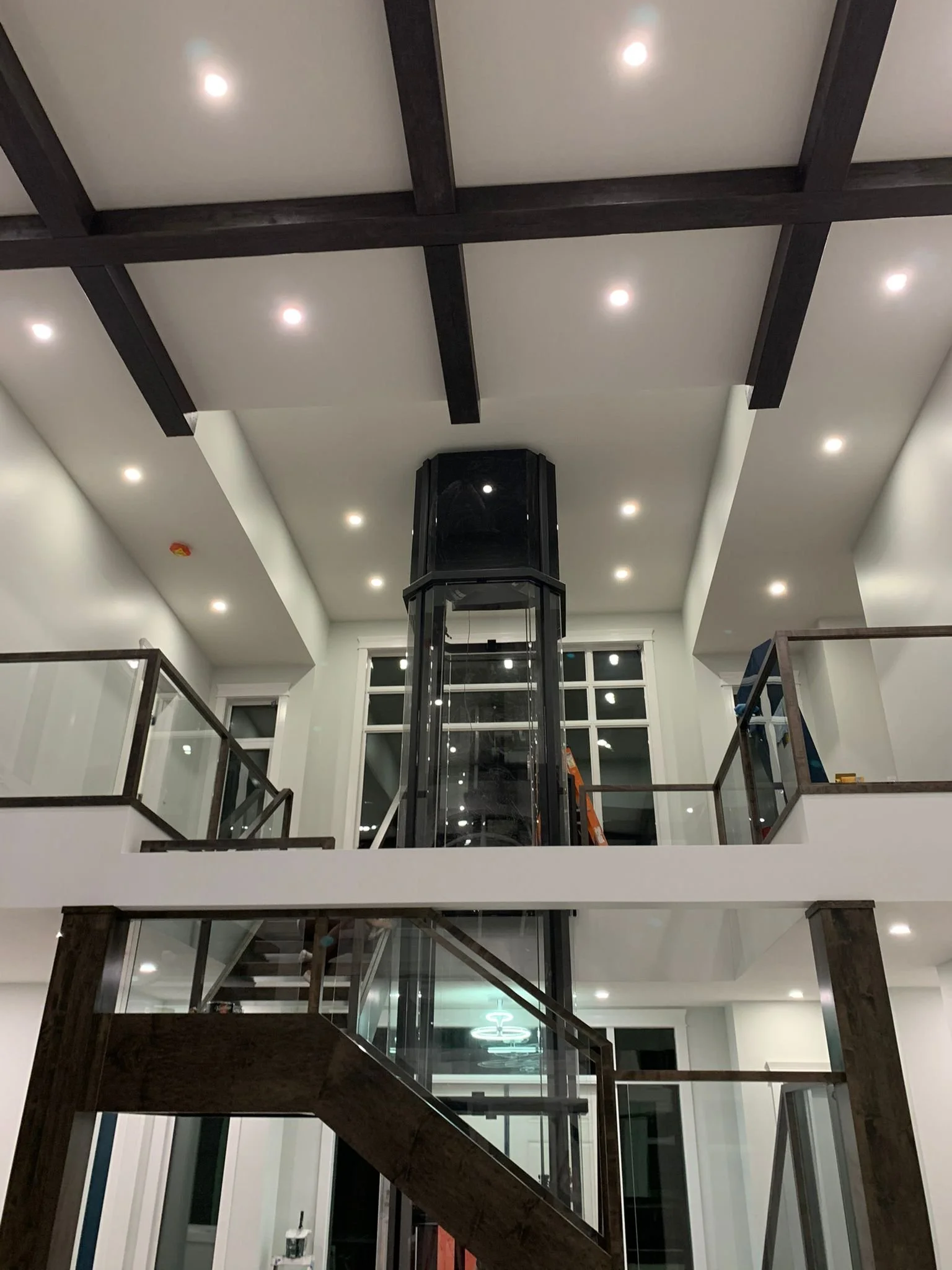 Interior view of a multi-story building with a modern elevator shaft in the center, surrounded by wooden staircase railings and ceiling lights.
