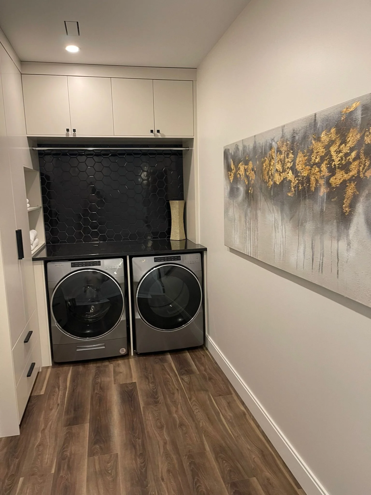 A laundry room with front-loading washer and dryer, white cabinets, a black hexagonal tile backsplash, and a large abstract painting on the wall.