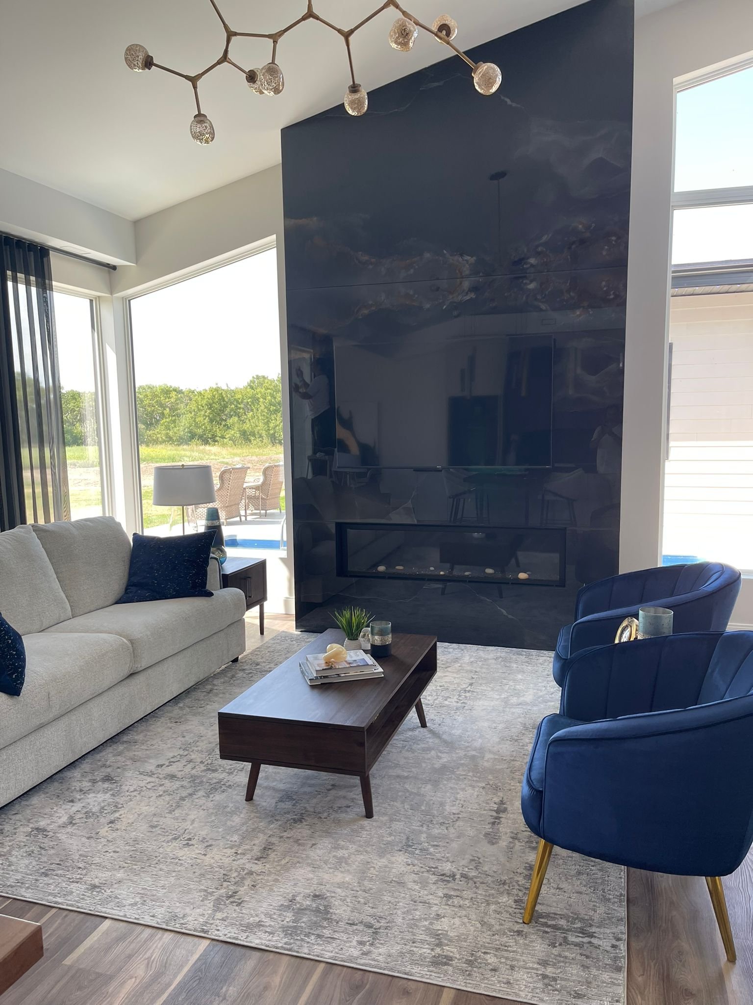 Living room with a beige sofa, blue armchair, coffee table, and a fireplace, with large windows showing outdoor patio and green landscape.