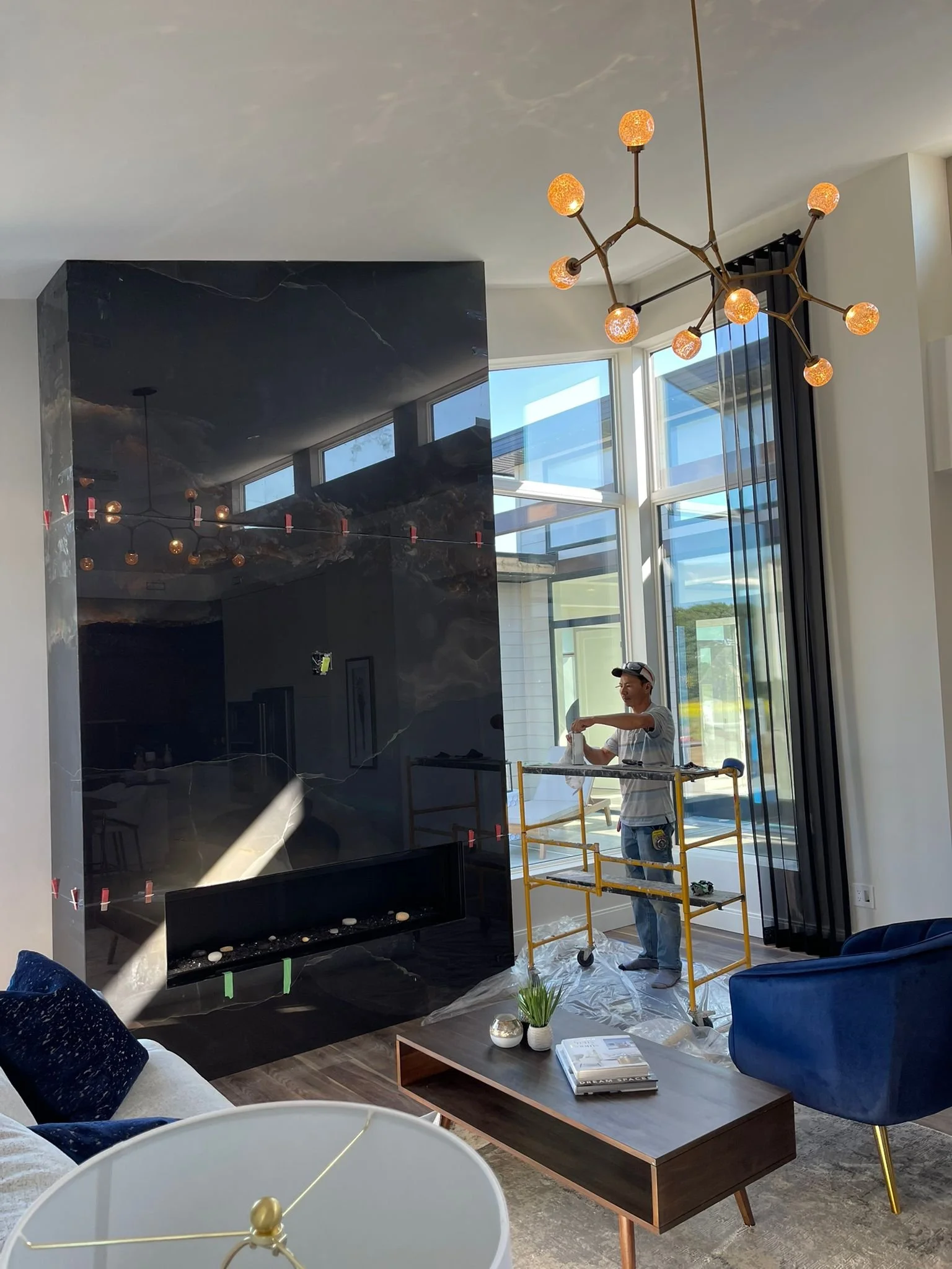 A living room with a modern black marble fireplace, a large window with black curtains, and contemporary furniture including a blue velvet armchair, a gray sofa with dark blue pillows, a wooden coffee table with magazines and decorative items, and a 