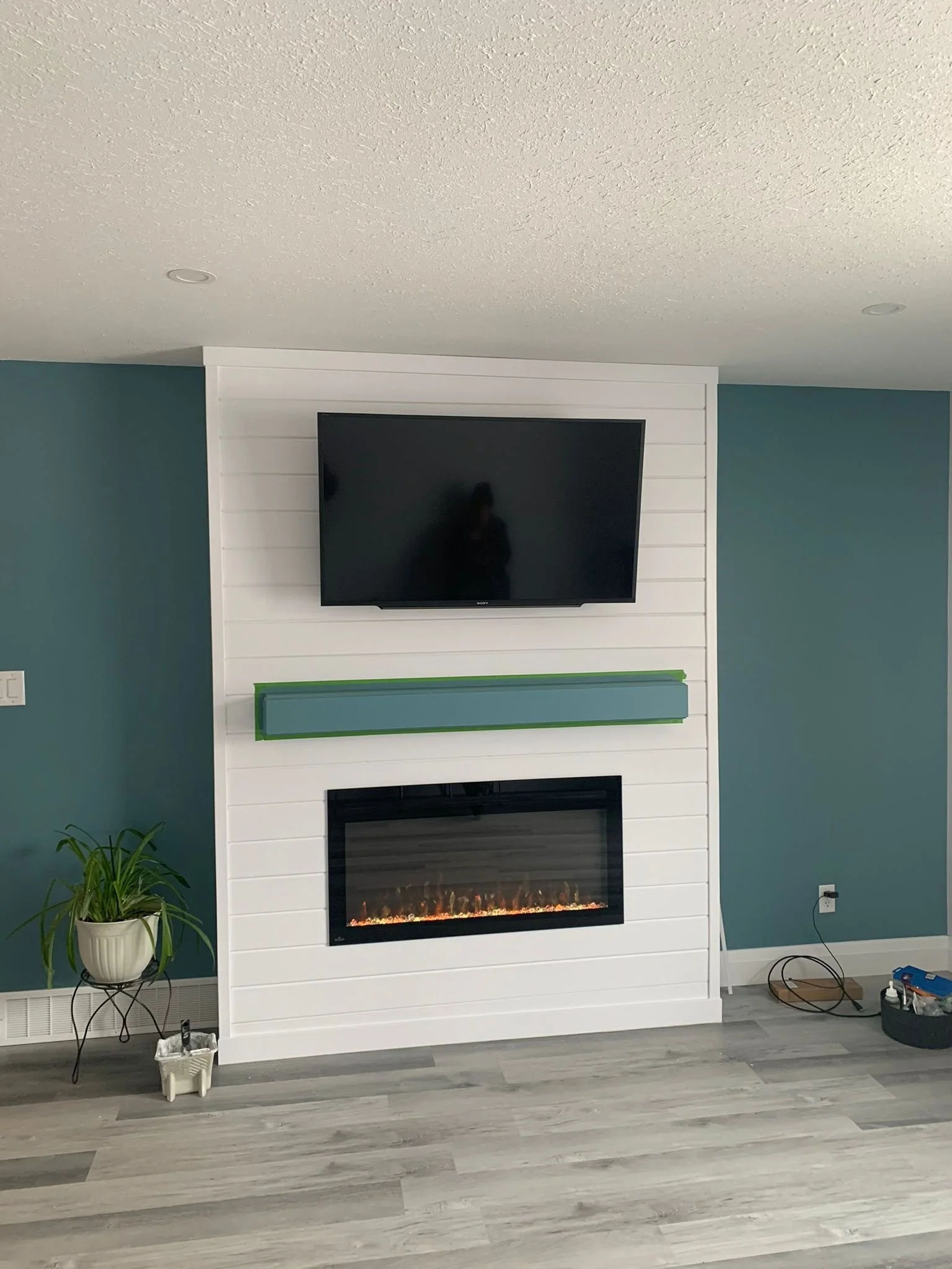 Living room with a flat-screen TV mounted on a white shiplap wall, electric fireplace below, teal accent wall on either side, potted plant on a small side table, hardwood flooring, and some electrical cords and tools on the floor.