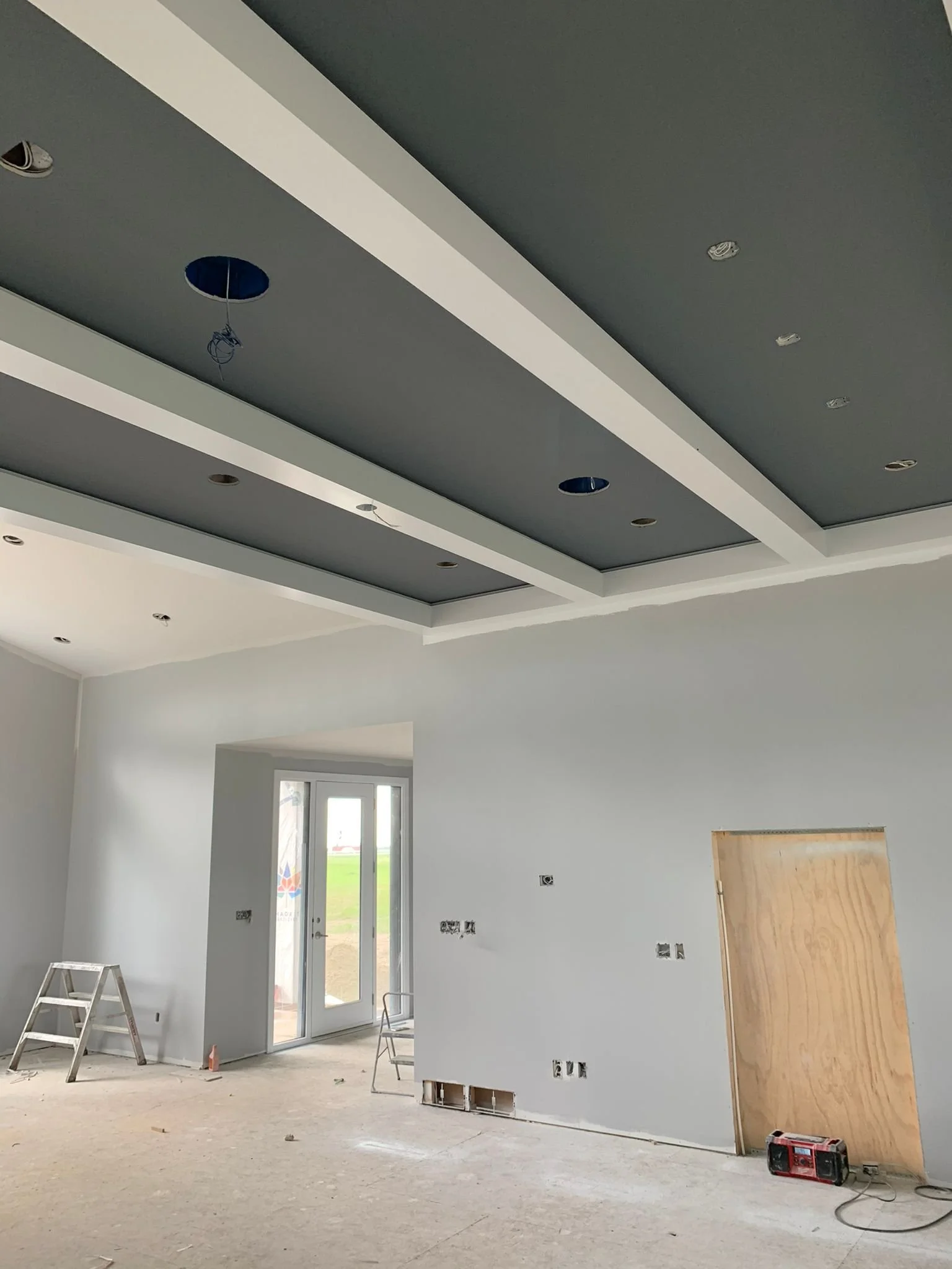 Interior of a house under construction, showing a room with a modern ceiling design, multiple electrical outlets and fixtures, and construction tools like a ladder, a wood panel, and a power drill on the unfinished floor.