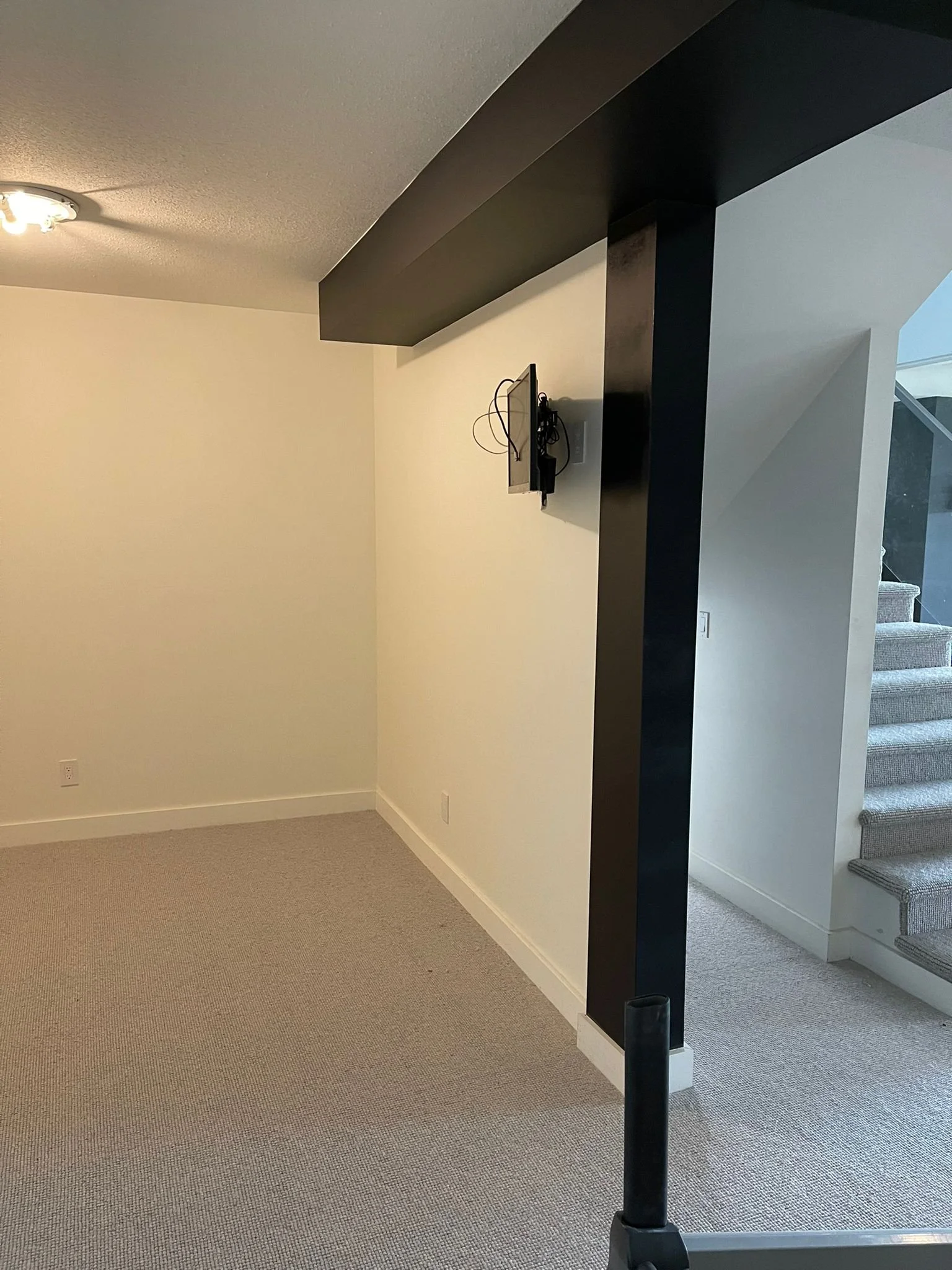 Empty room with beige carpet, cream-colored walls, a black beam extending from the ceiling to the wall, and stairs leading upward in the background.