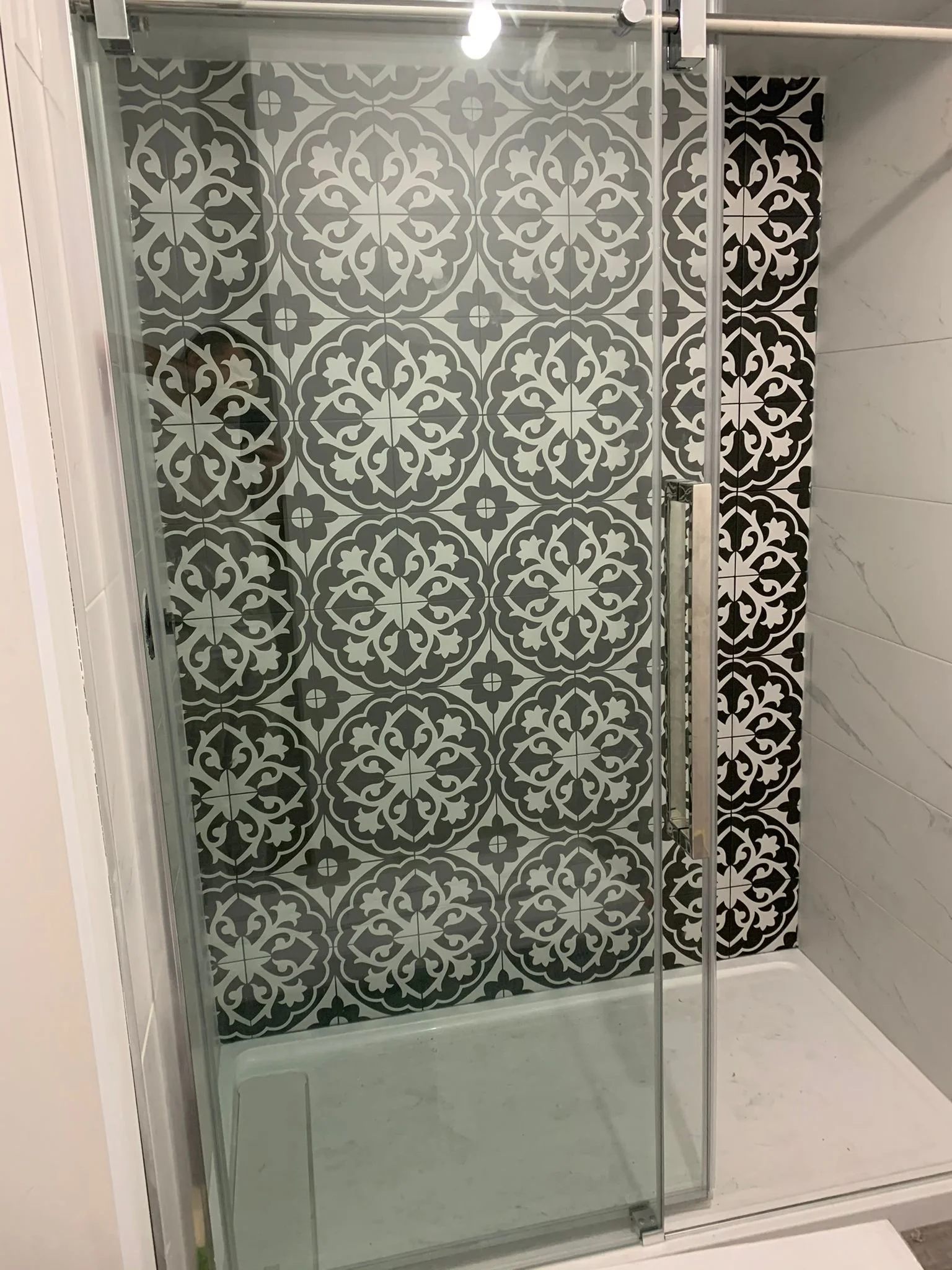A glass shower enclosure with black and white patterned tiles on the back wall and a white marble tile on the right side. The shower has a sliding glass door with a metal handle.