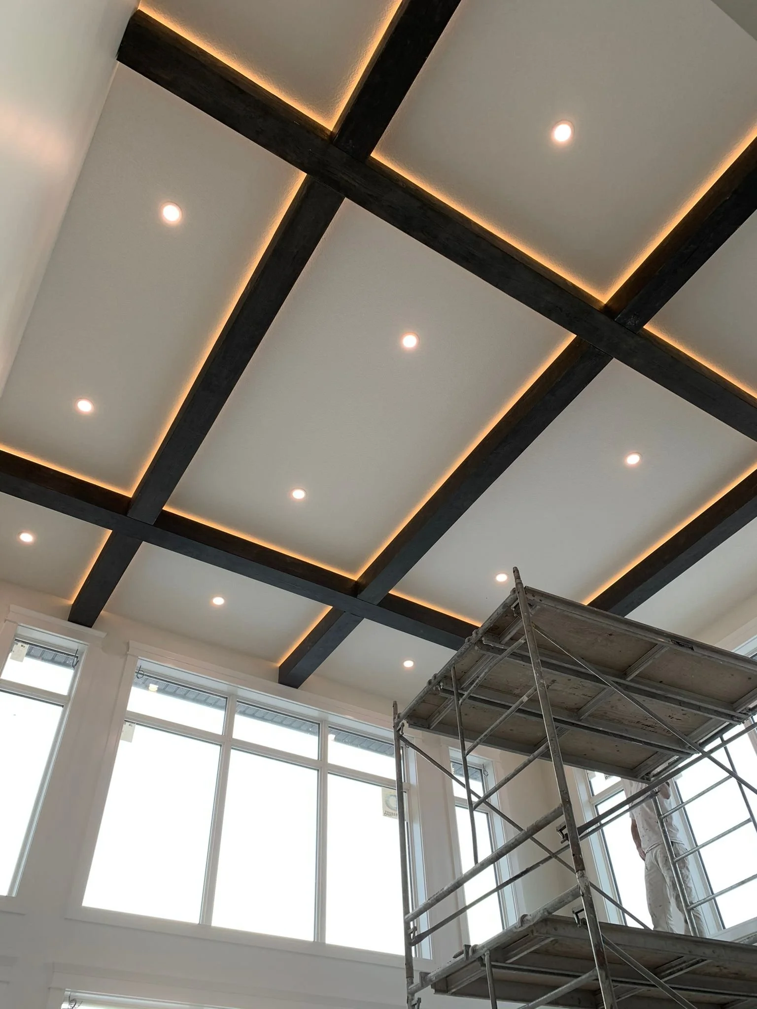 Interior ceiling with dark wooden beams and recessed lighting, large windows allowing natural light, scaffolding and worker present indicating ongoing renovation or construction.