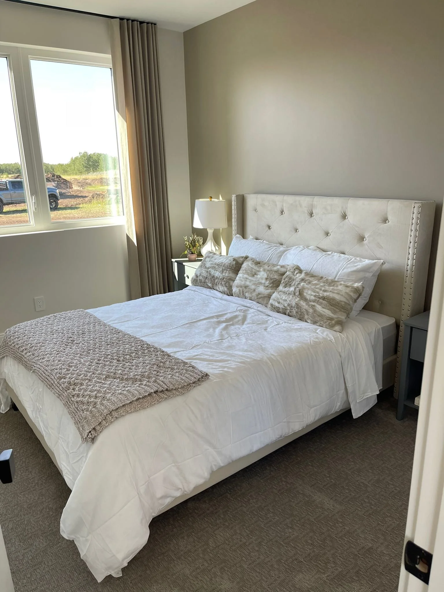 A neatly made bed with white bedding and pillows, a beige fur throw on top, a nightstand with a white lamp, a small plant, and window with beige curtains letting in natural light.