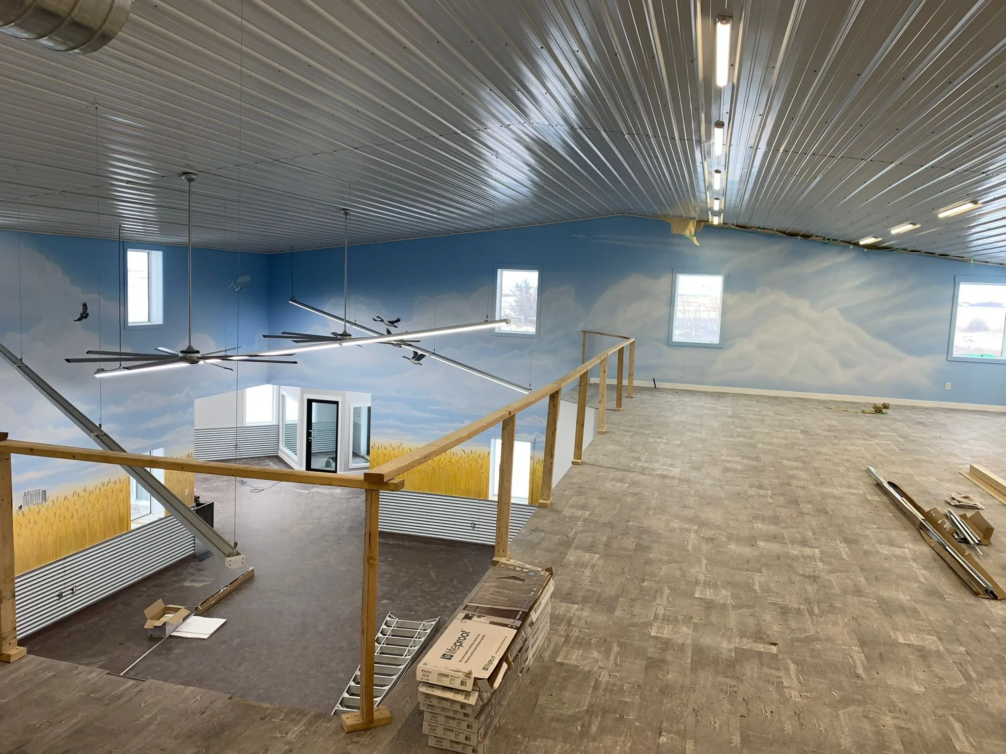 Construction site interior with a newly painted blue sky and cloud mural, a wooden railing, and hanging light fixtures. Construction materials and tools are scattered on the floor.