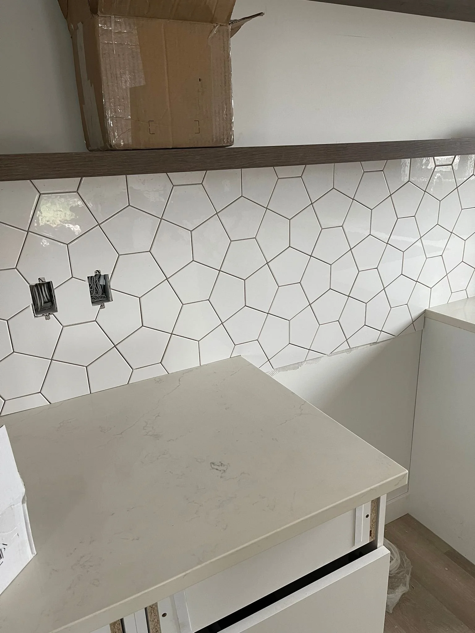 Interior of a space under construction or renovation showing a section of a tiled wall with hexagon-shaped white tiles. There are two electrical outlets missing cover plates, and a white countertop or shelf with some scratches. A cardboard box is pla