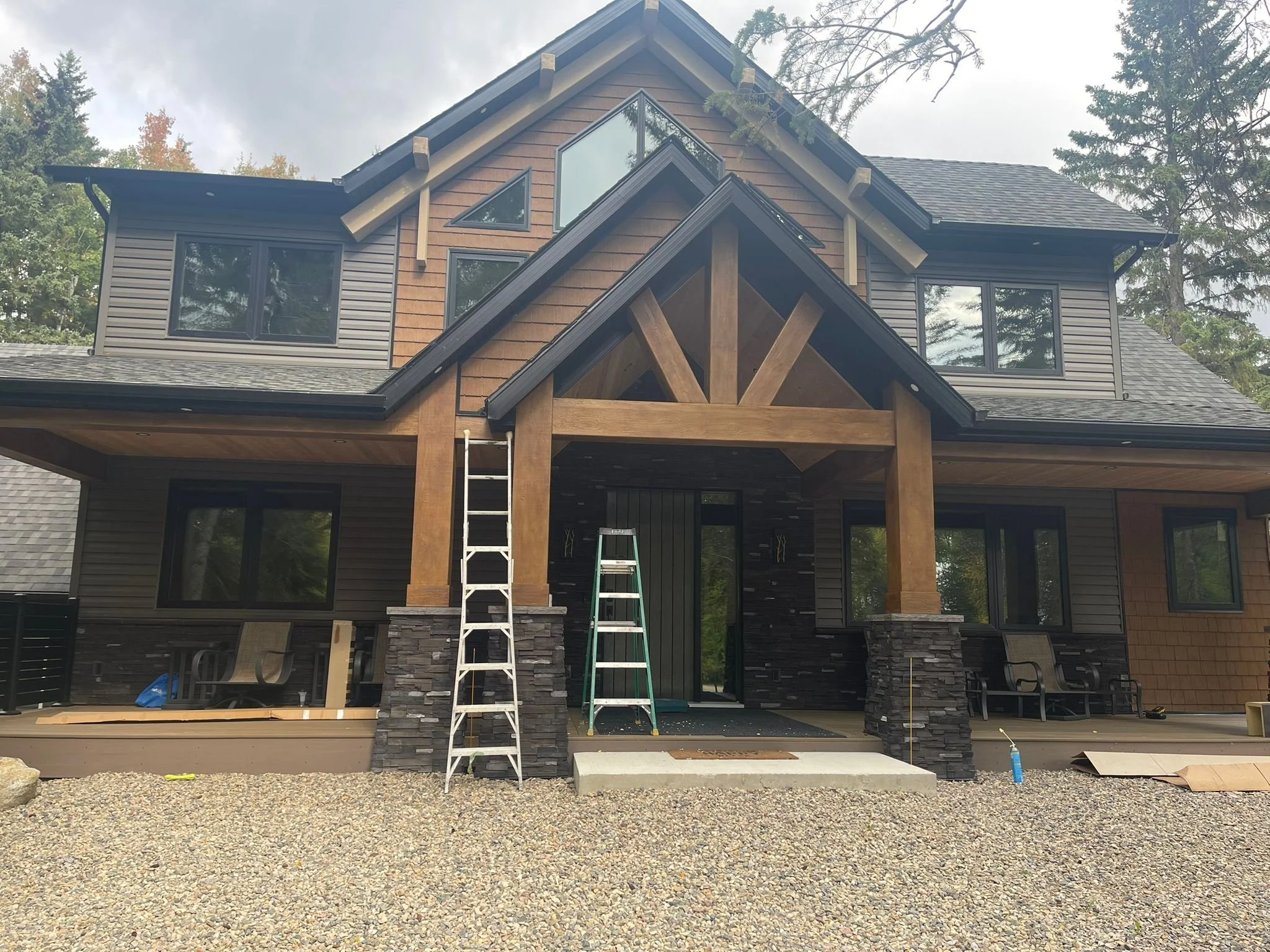 A house under construction with wooden beams and stone accents, with two ladders and construction materials in the foreground and trees in the background.