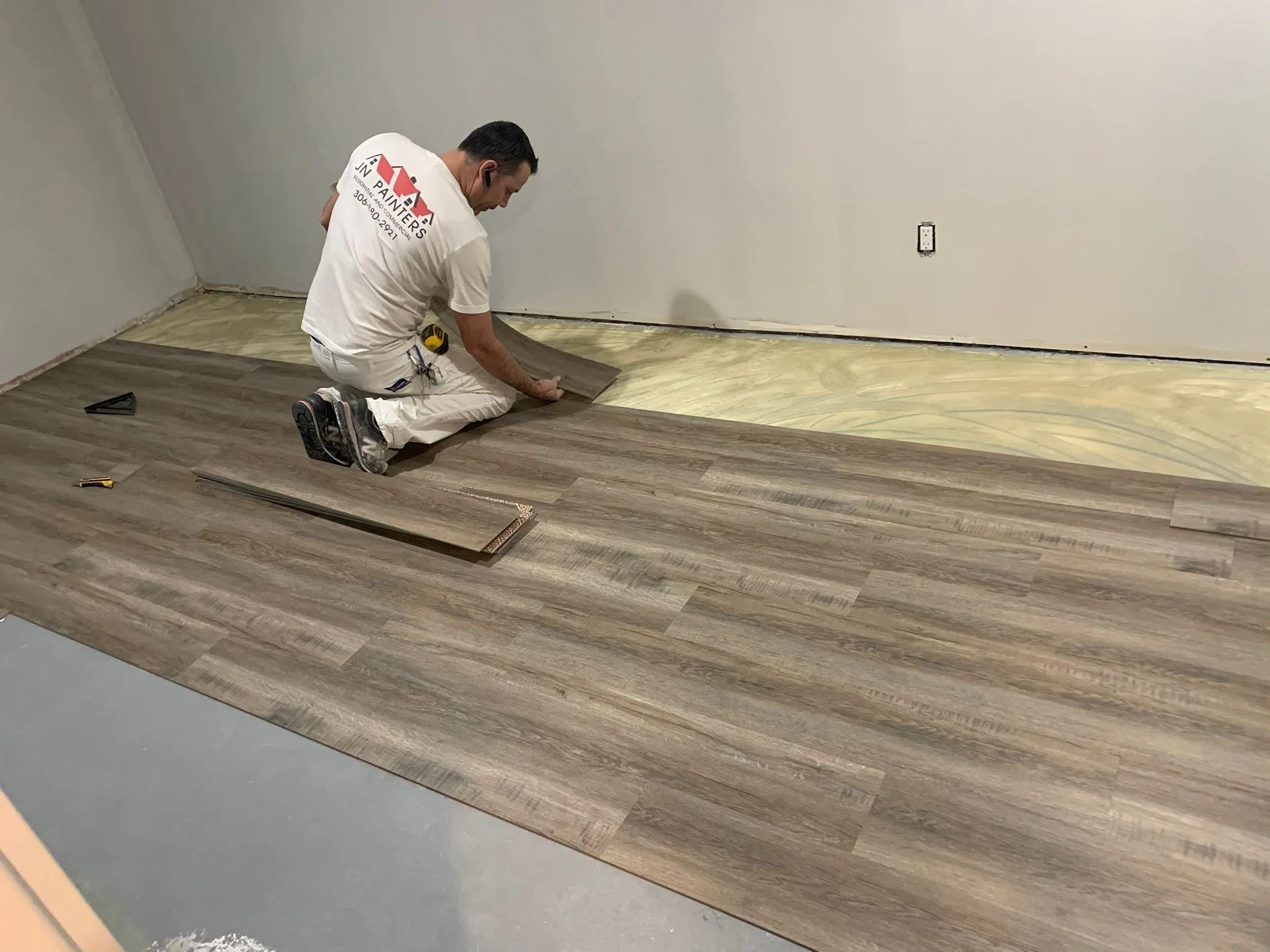 A man kneeling on the floor installing wood or laminate flooring in a room with plain white walls. He is wearing a white t-shirt with text on the back and is handling a flooring plank with tools nearby.
