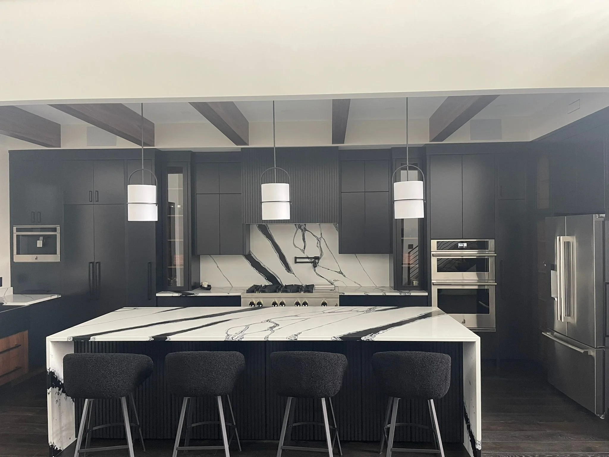 Modern kitchen with black cabinets, white marble island, three pendant lights, stainless steel appliances, and dark wood ceiling beams.