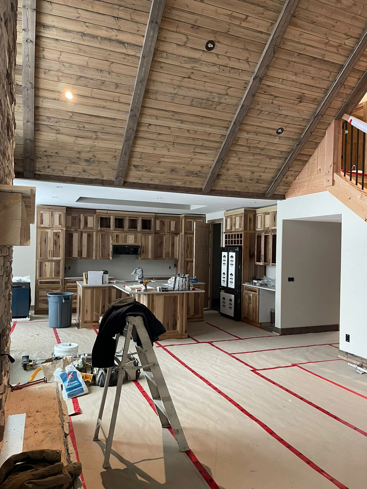 Interior of a house under construction, with a kitchen area featuring wooden cabinets, a countertop, and appliances. The ceiling is made of wood with exposed beams, and the floor is covered with protective paper marked with red lines. Construction to