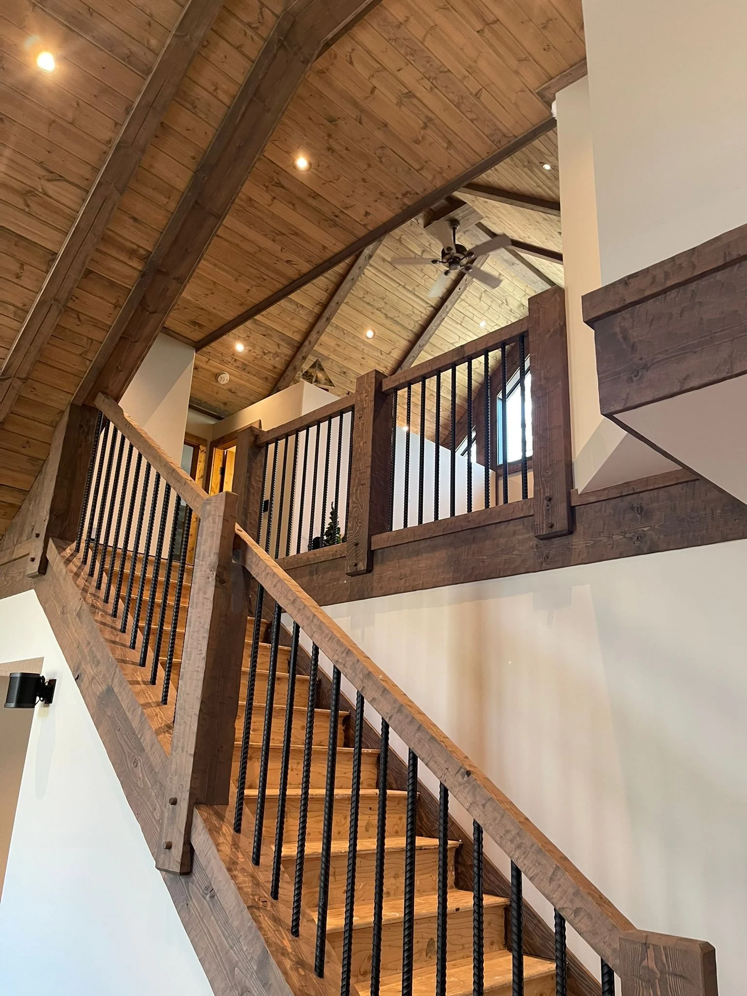 Interior view of a wooden staircase with black metal balusters leading up to a second floor with a wooden ceiling, ceiling fan, and windows.